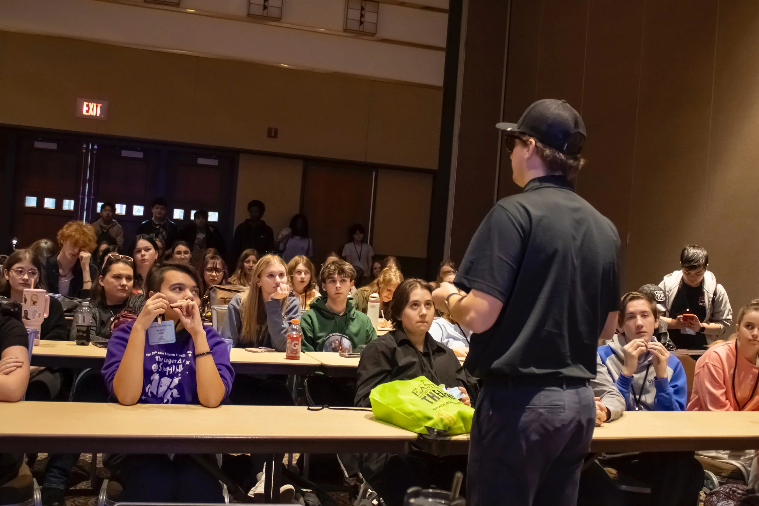 A speaker, wearing a black cap, black shirt, and glasses, stands addressing a diverse group of teenagers seated at long tables inside a conference room or auditorium. The young audience listens attentively, some with notebooks and drinks in front of them.
