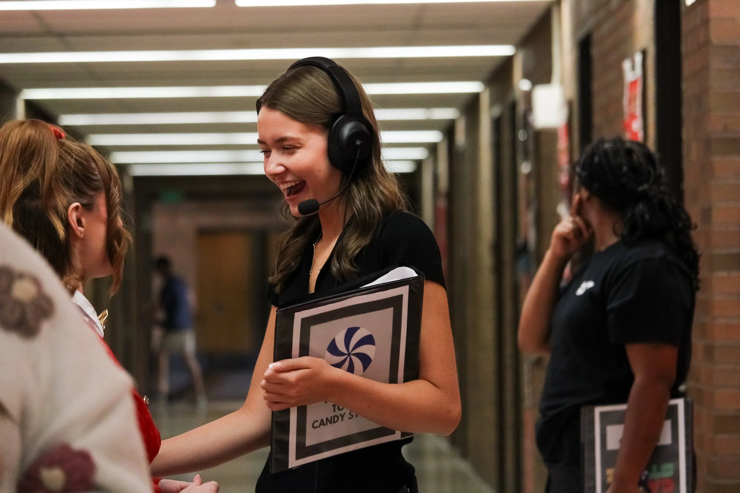 A woman with a headset and clipboard smiling and talking to a young girl in a hallway, with another woman standing nearby.