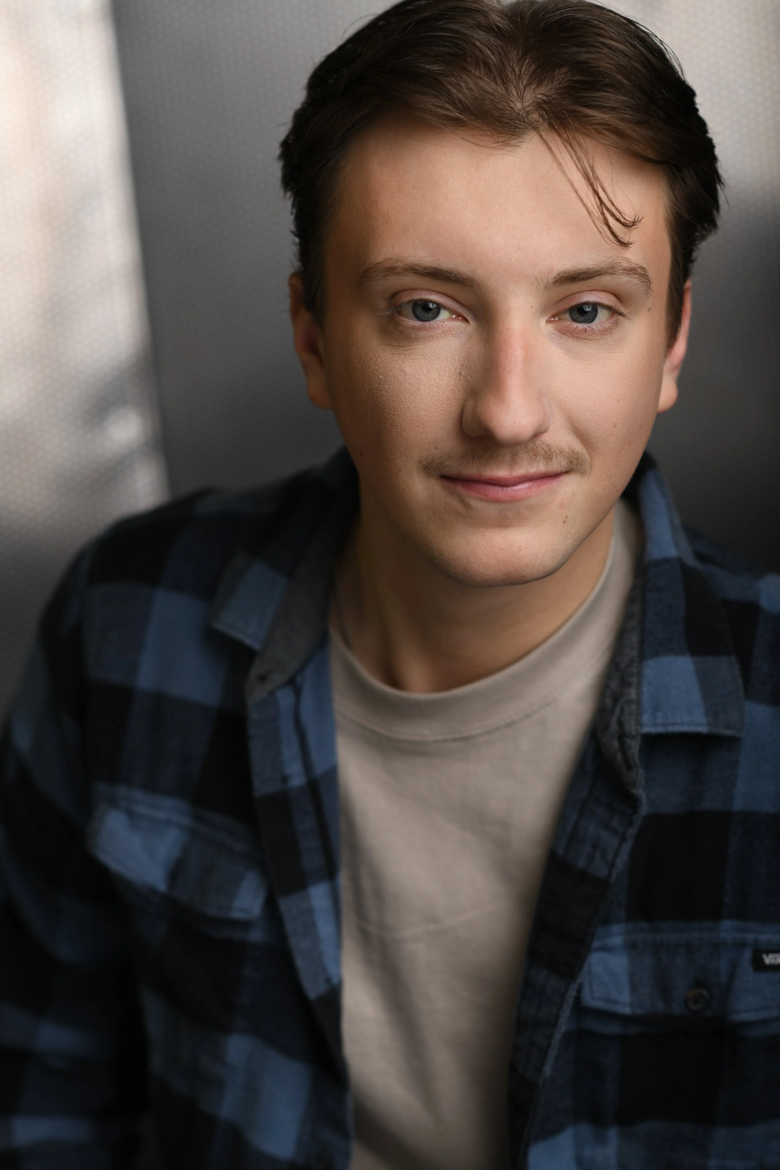 A young man with light brown hair, blue eyes, and a slight smile, wearing a black and blue checkered shirt over a beige t-shirt.