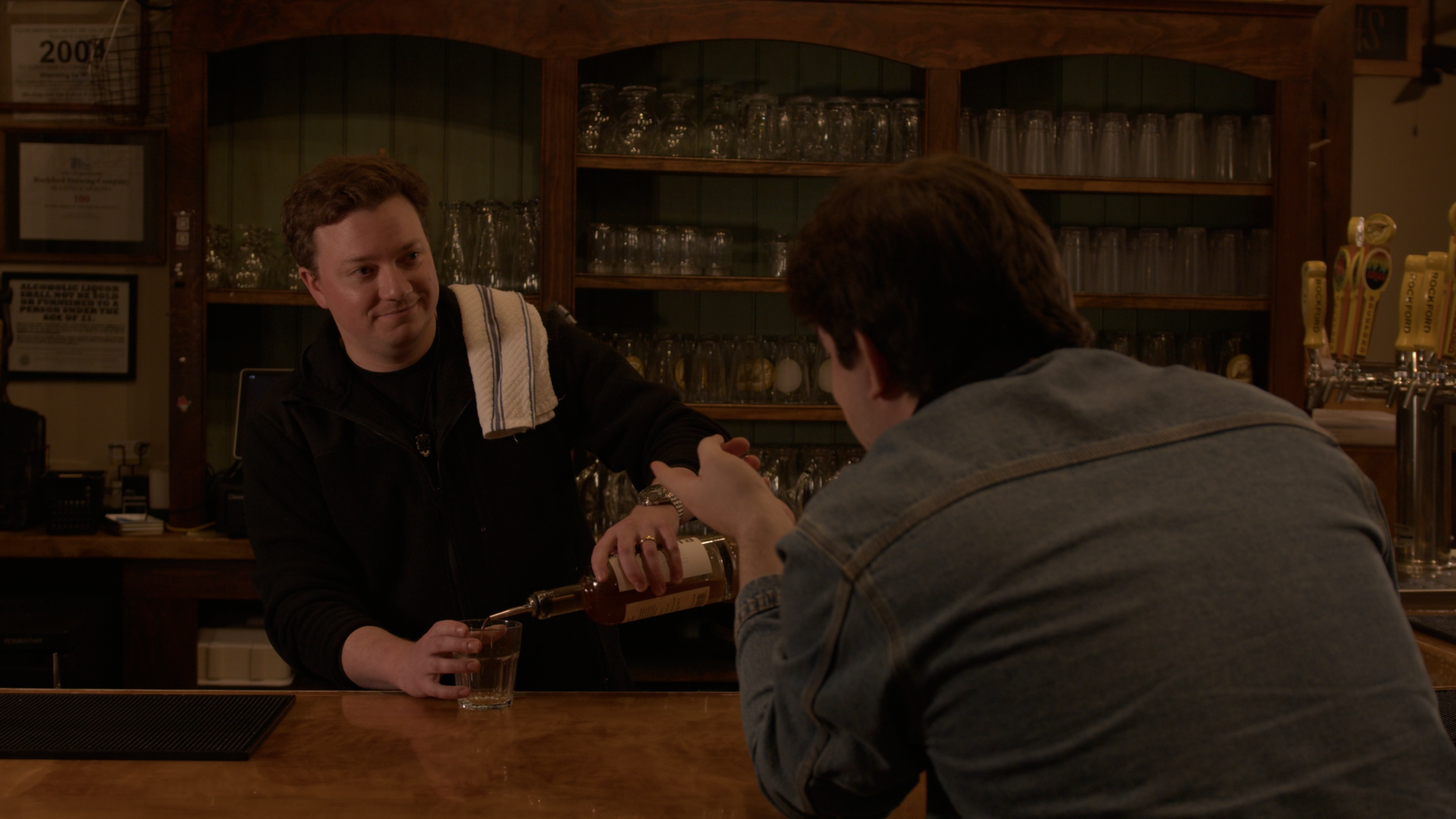 A bartender pours whiskey into a glass for a customer at a bar. The bartender has a towel over his shoulder and is smiling, while the customer is leaning on the bar with his hands together.