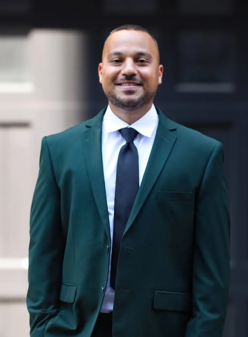 A smiling man in a dark green suit, white shirt, and black tie standing outdoors in front of a modern building.