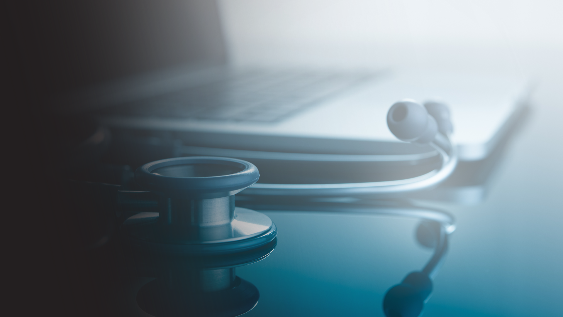 Close-up of a stethoscope placed on a reflective medical table near a laptop keyboard in a medical or clinical setting.
