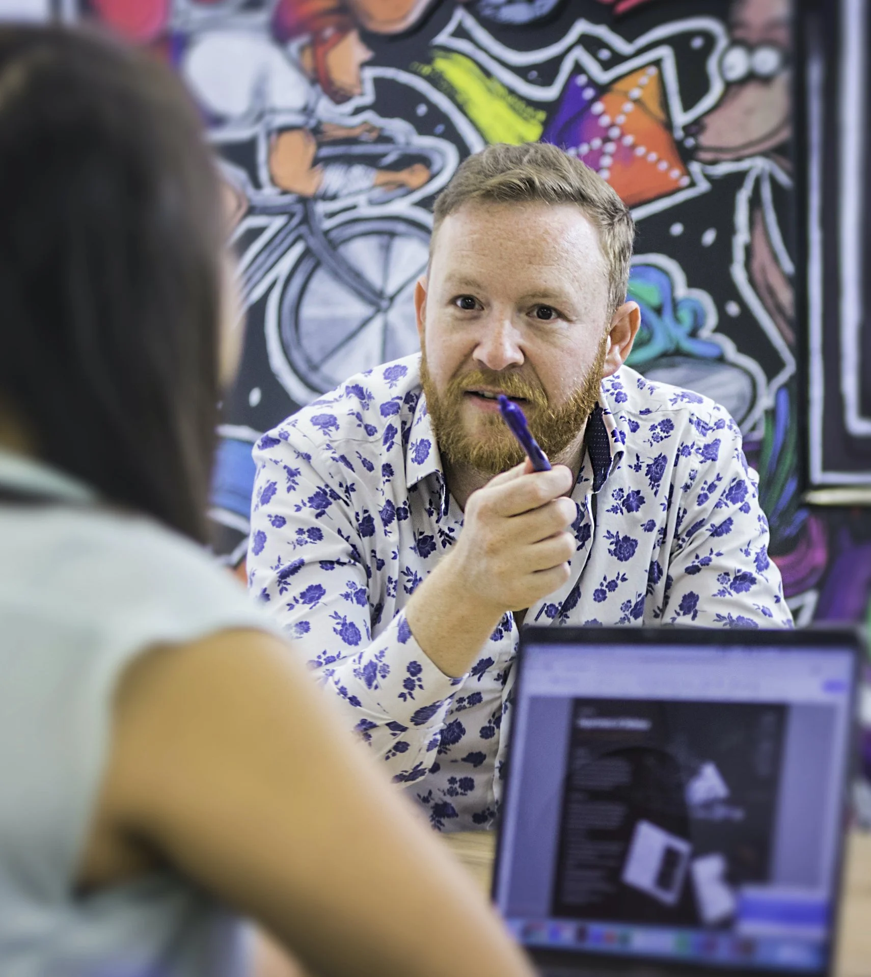 Jason, wearing a white shirt with blue floral patterns, holding a pen while engaging in a conversation. He is seated in front of a colourful mural featuring a cyclist.
