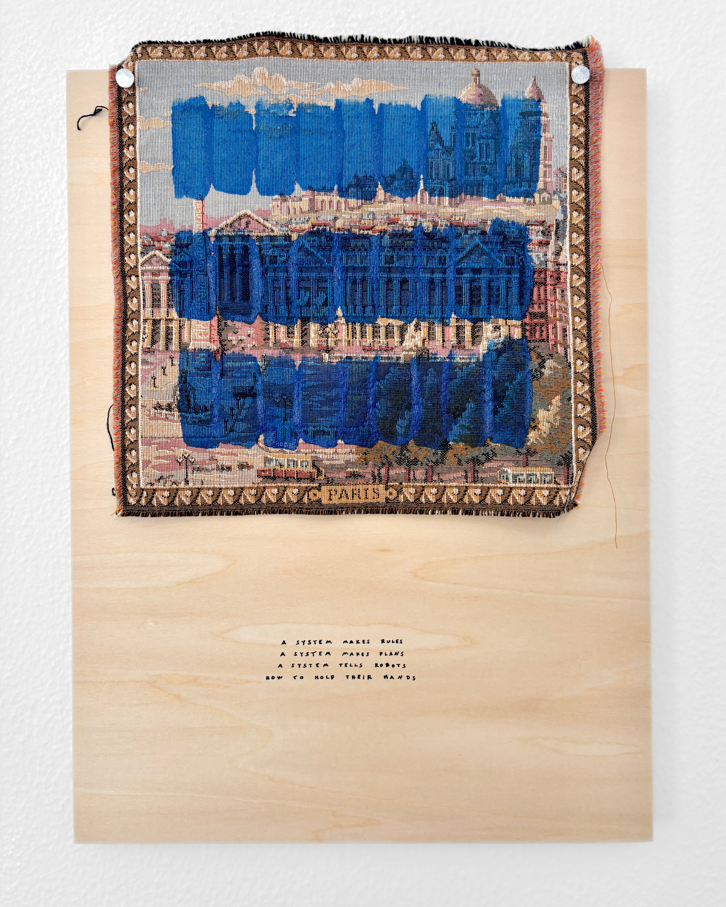 Painted textile mounted on a wooden board with handwritten text underneath reading: 'A system makes rules, a system makes plans, a system tells robots how to hold their hands.' Original artwork by Philip Hauge Reitz.