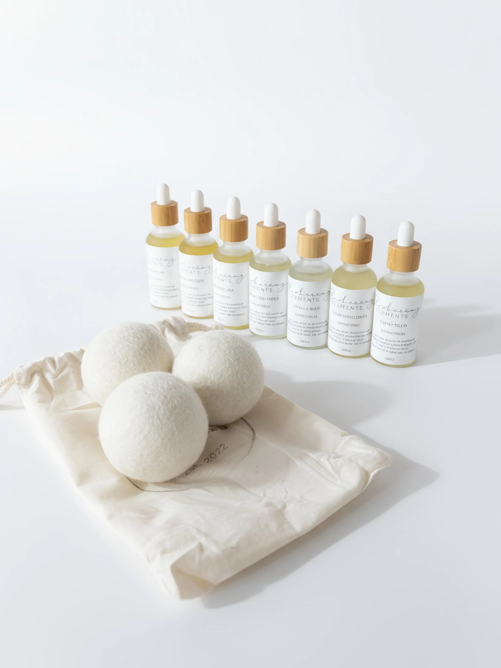 Collection of scented oils including seven small glass bottles with droppers and white labels, a beige drawstring bag, and three white clothes dryer balls on a light-colored surface.