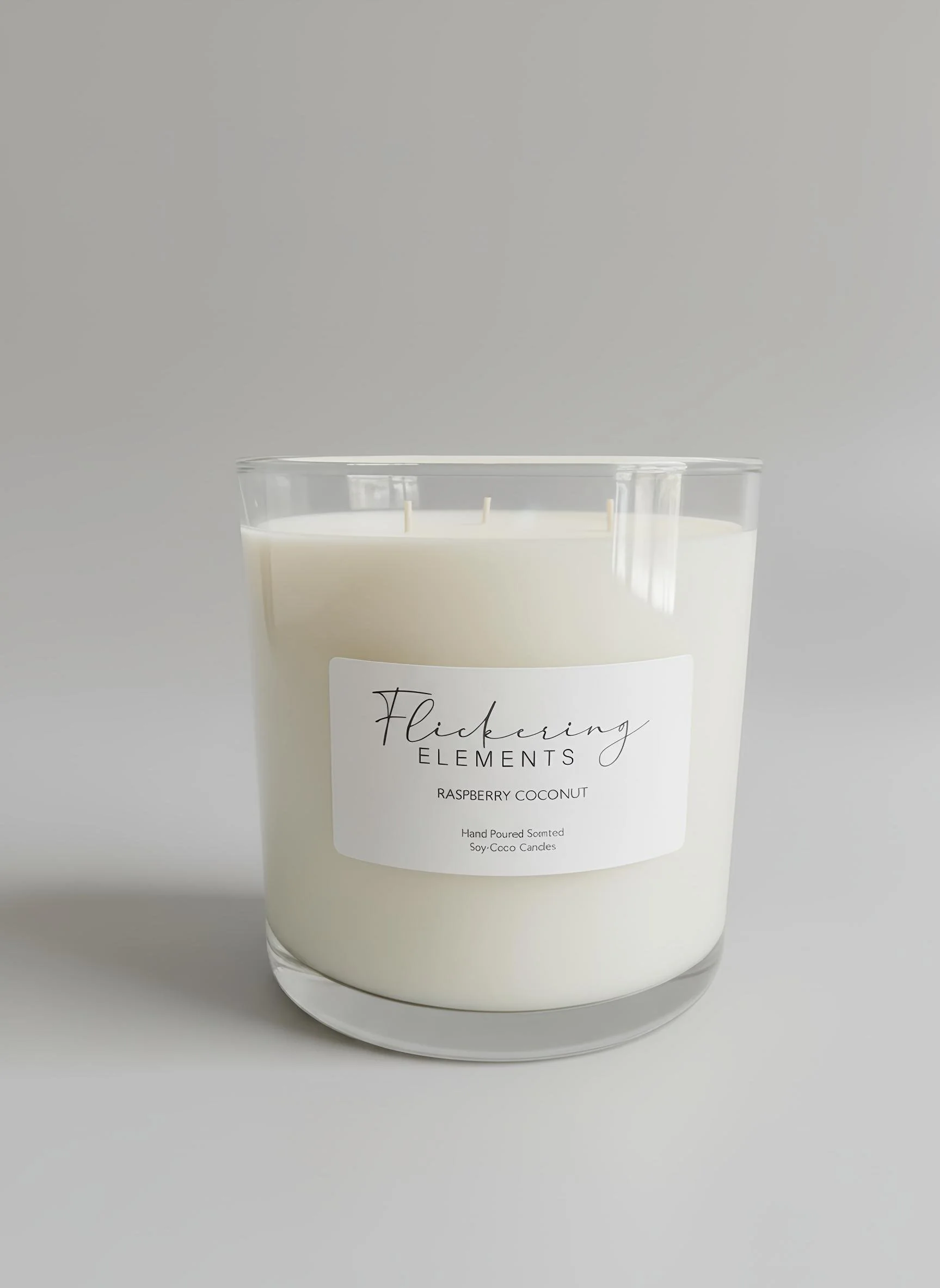A white scented candle in a clear glass container with a label reading "Flickering Elements Raspberry Coconut" on a plain light gray background.