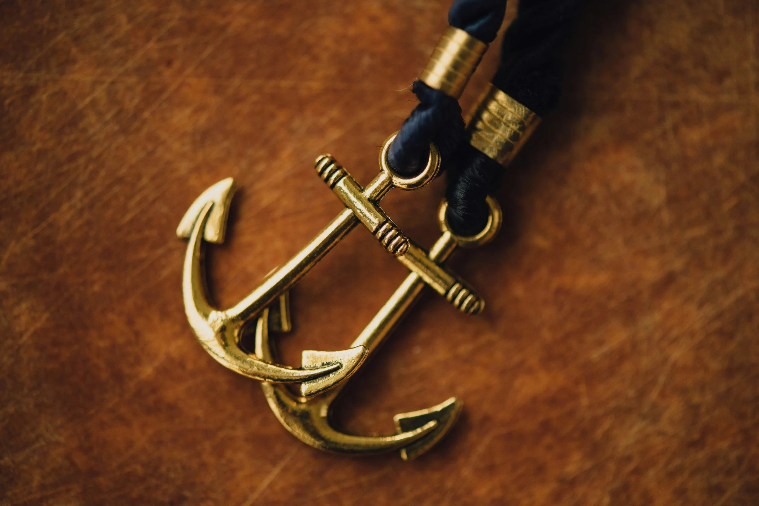 Close-up of two gold anchor-shaped cufflinks with black fabric and gold accents, placed on a wooden surface.