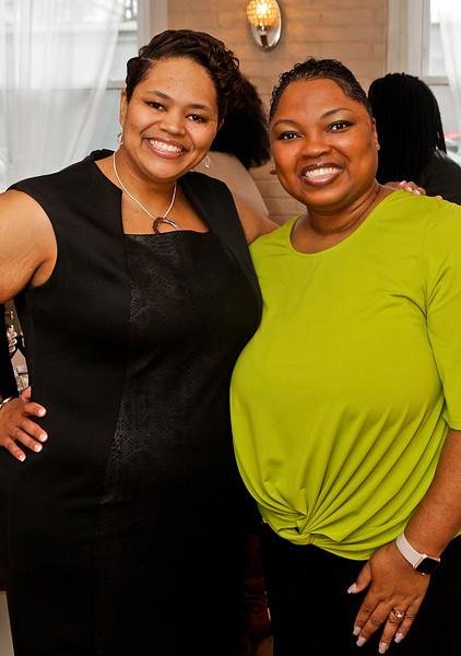 Two women smiling and posing for a photo indoors, one in a black dress, the other in a bright green top.