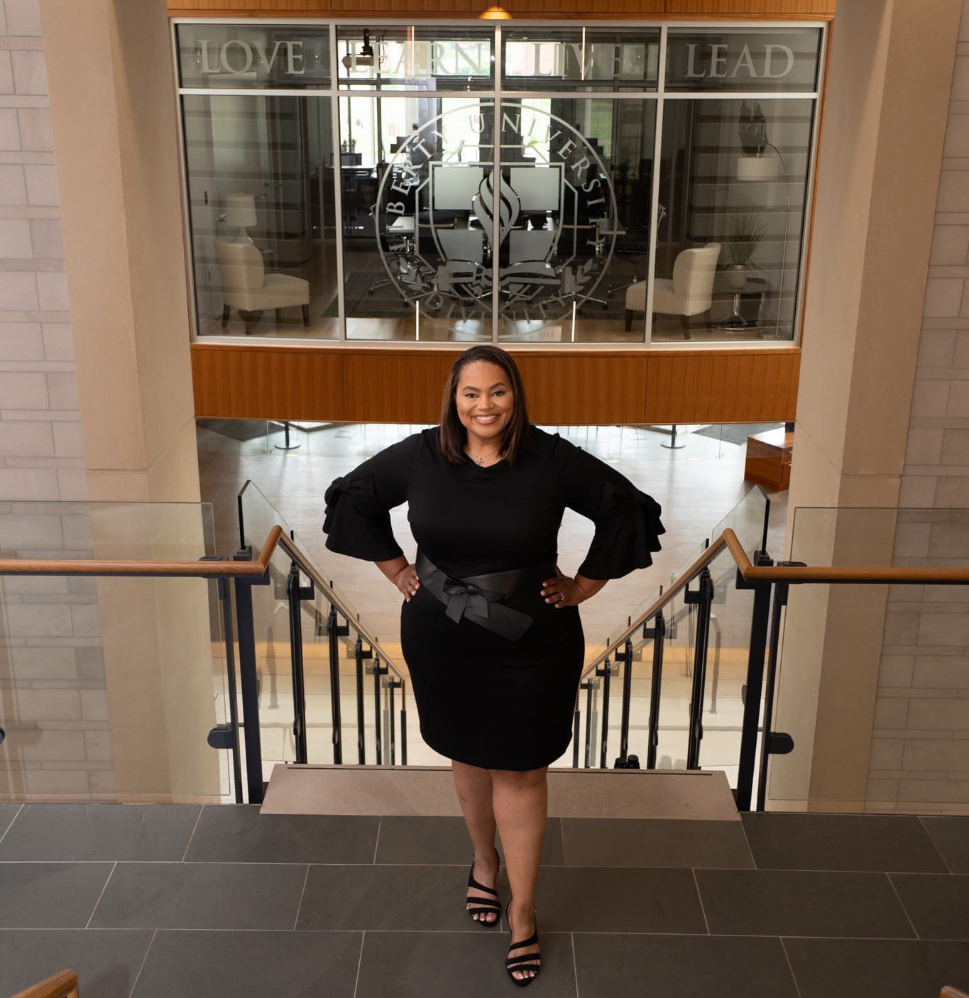A woman in a black dress with a belt, standing on a staircase inside a building, smiling at the camera. Behind her is a glass-walled office with the university logo and the words 'Love,' 'Learn,' and 'Lead' displayed at the top.