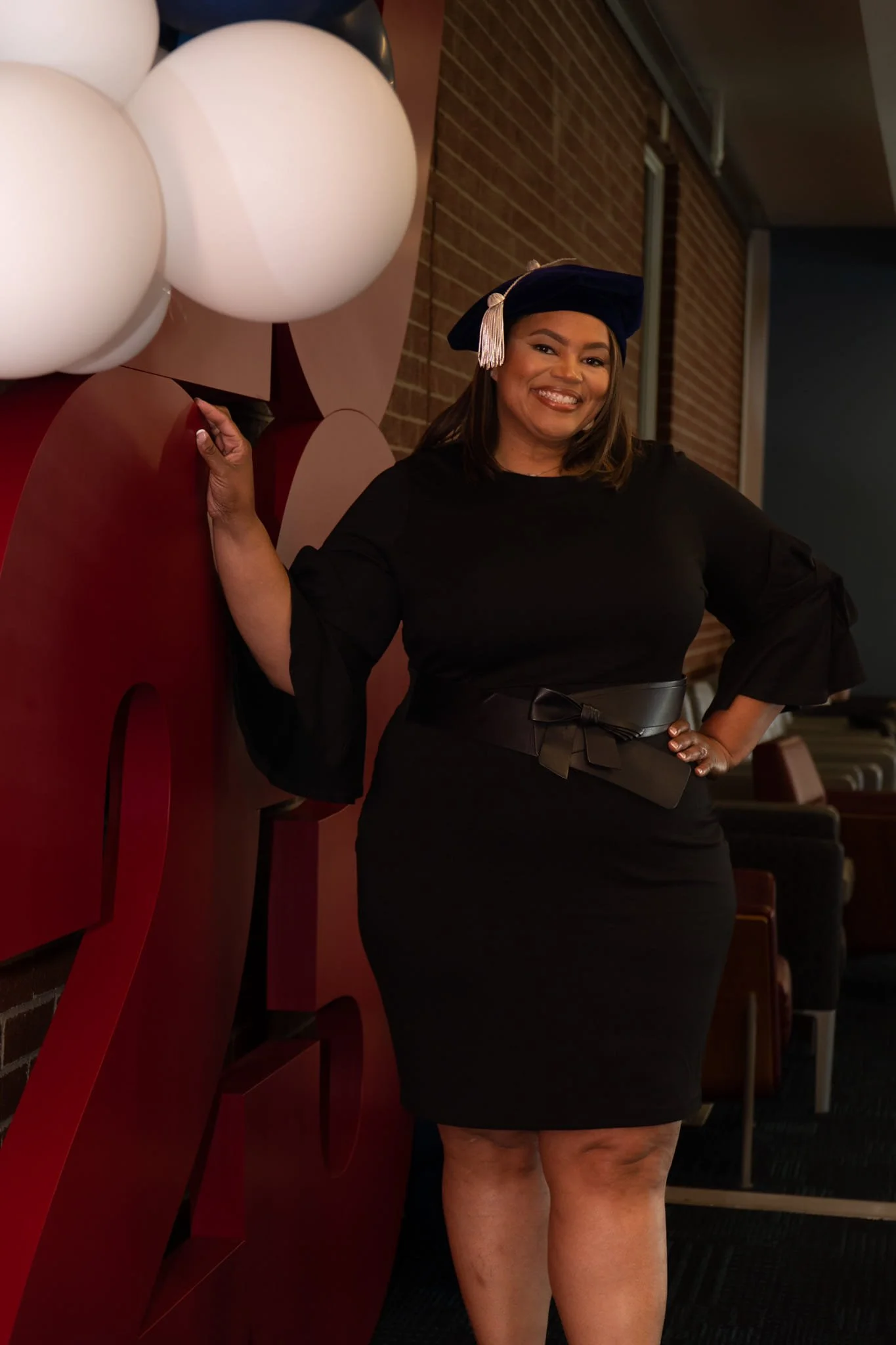 Woman wearing a graduation cap and black dress, smiling and standing next to large decorative balloons.