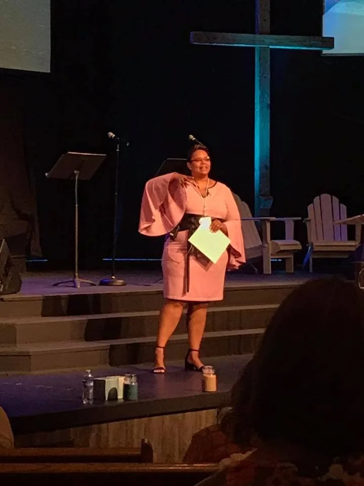 A woman in a pink dress stands on stage holding papers, smiling, with a large wooden cross behind her. There are music stands and chairs on stage, and audience members are visible in the foreground.