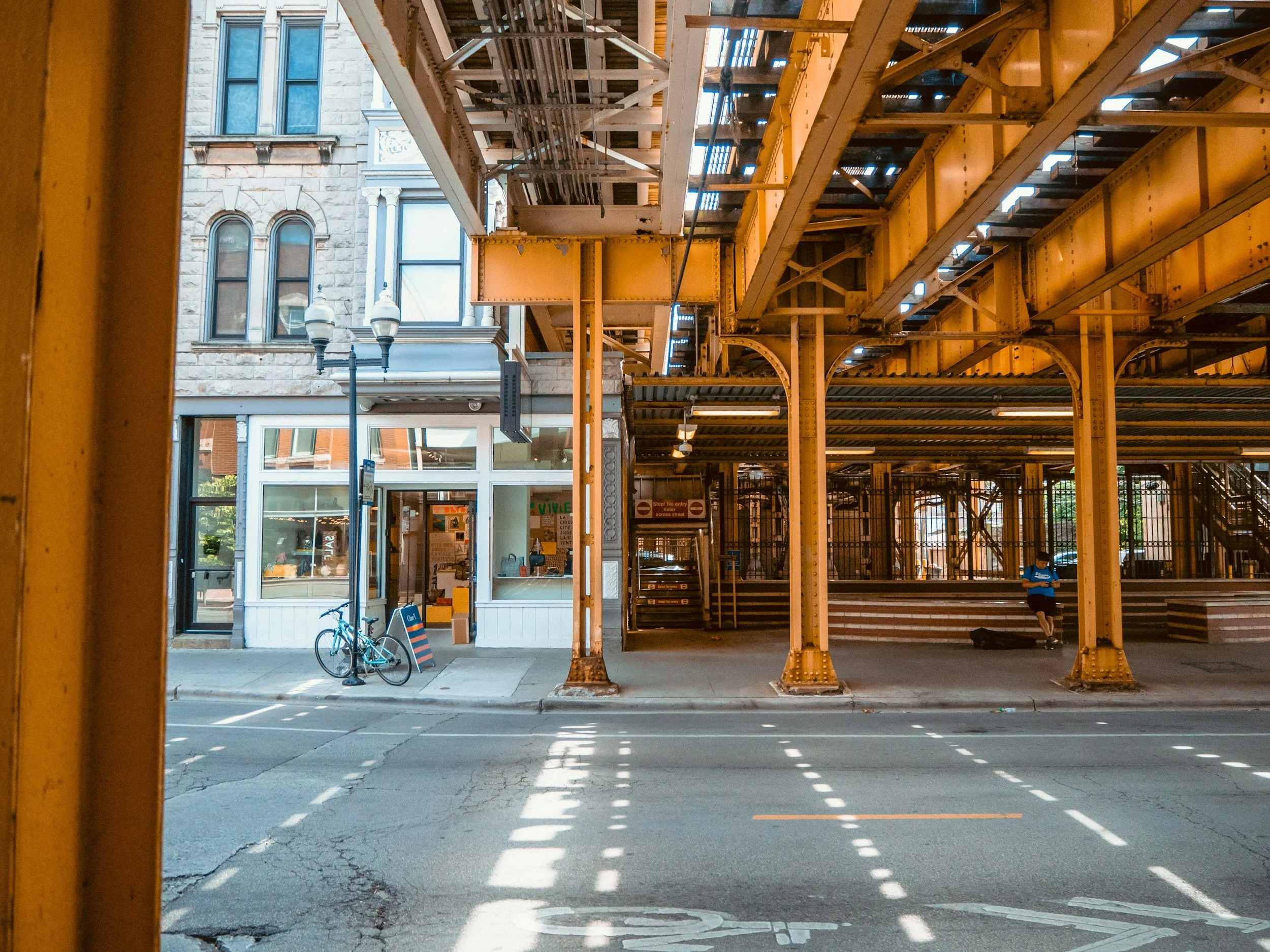 View of a city street underneath an elevated train track with a small storefront and a person sitting on a bench.