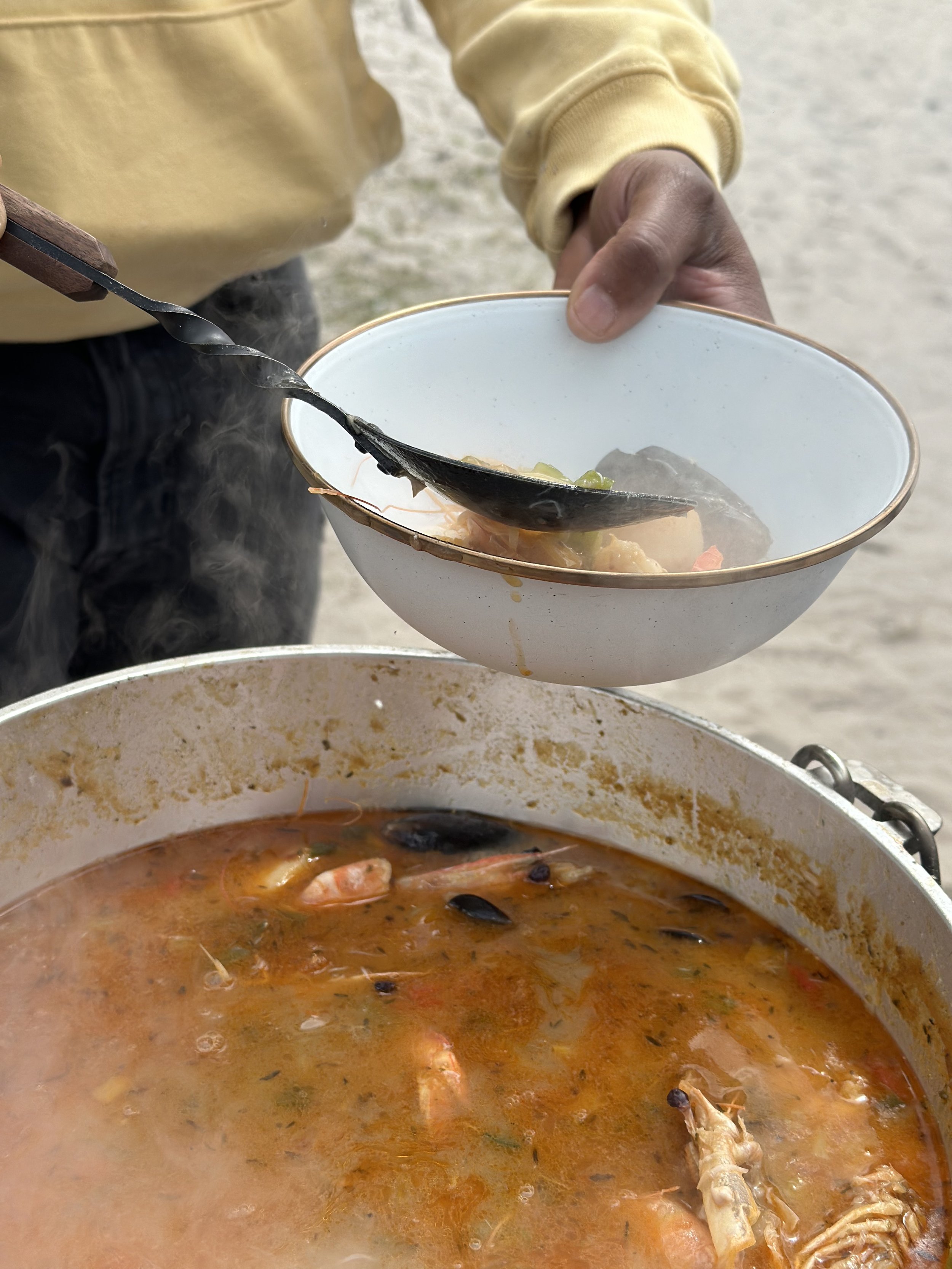 Person serving hot seafood soup from a large pot into a bowl outdoors.
