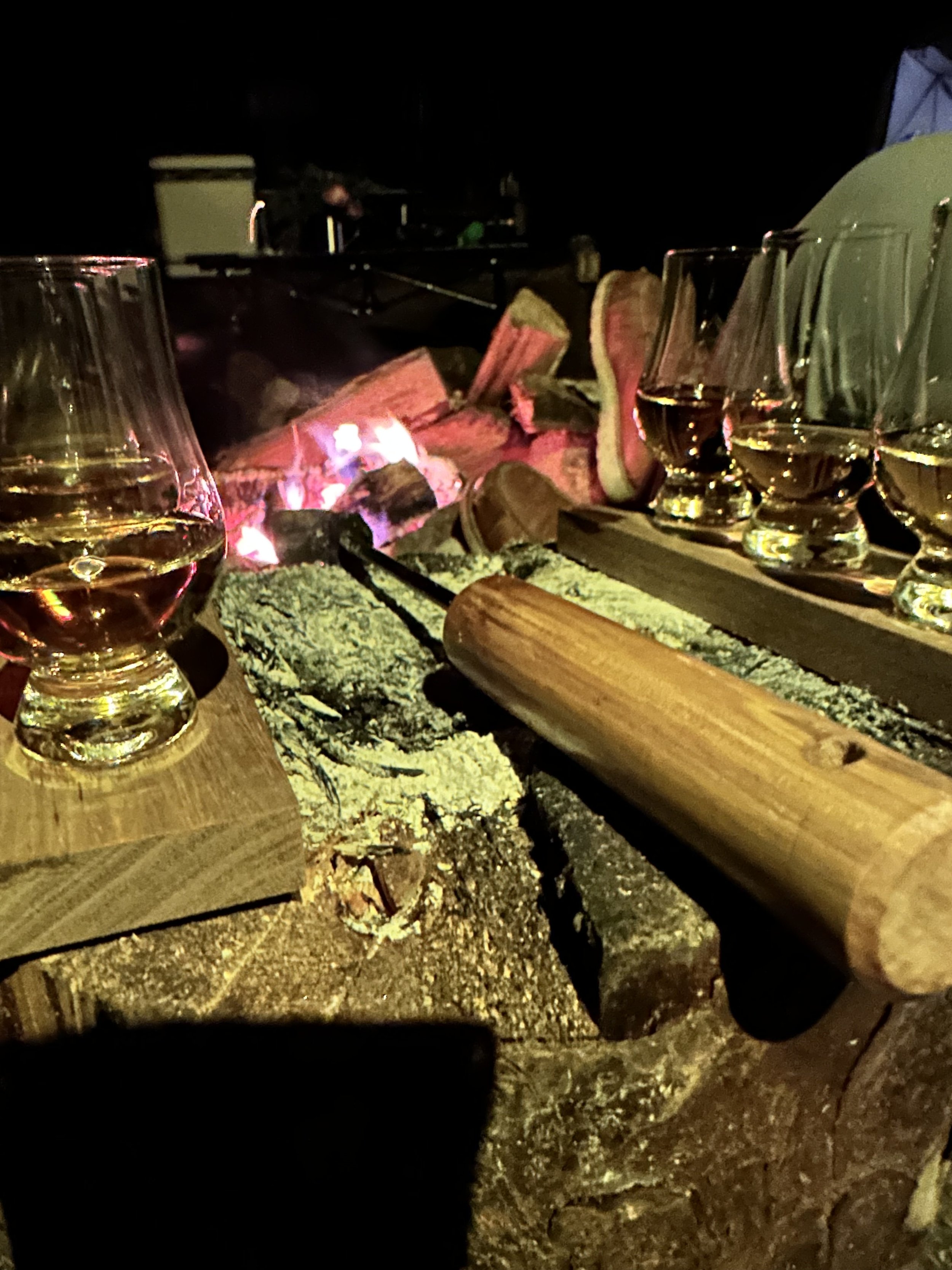 Four glasses of white wine on wooden coasters around a crackling fire with logs and a fire poker, in a dark outdoor setting.