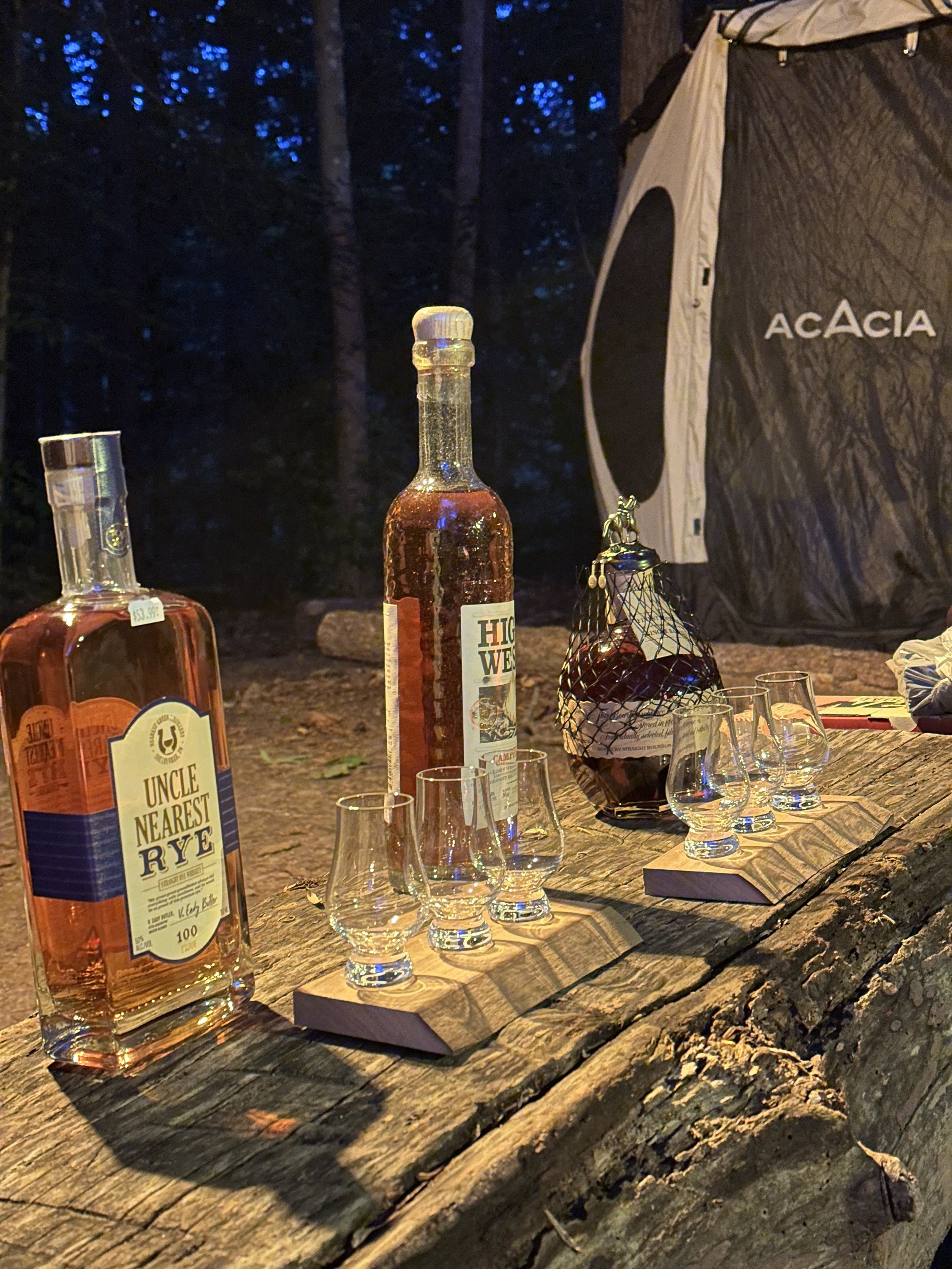 Charcoal-colored tent with the brand name 'Acacia', set up outdoors at dusk or evening. In front of the tent is a rustic wooden table holding bottles of whiskey, one labeled 'Uncle Nearest Rye' and another with a different label, along with small tasting glasses on wooden holders and a bag of ice or dark liquid, likely whiskey or bourbon. The background features dark blue sky and tall trees.