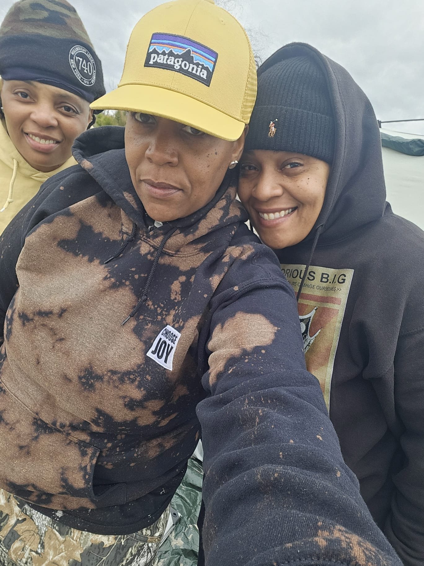 Three friends taking a selfie outdoors, dressed in casual and warm clothing, with cloudy skies in the background.