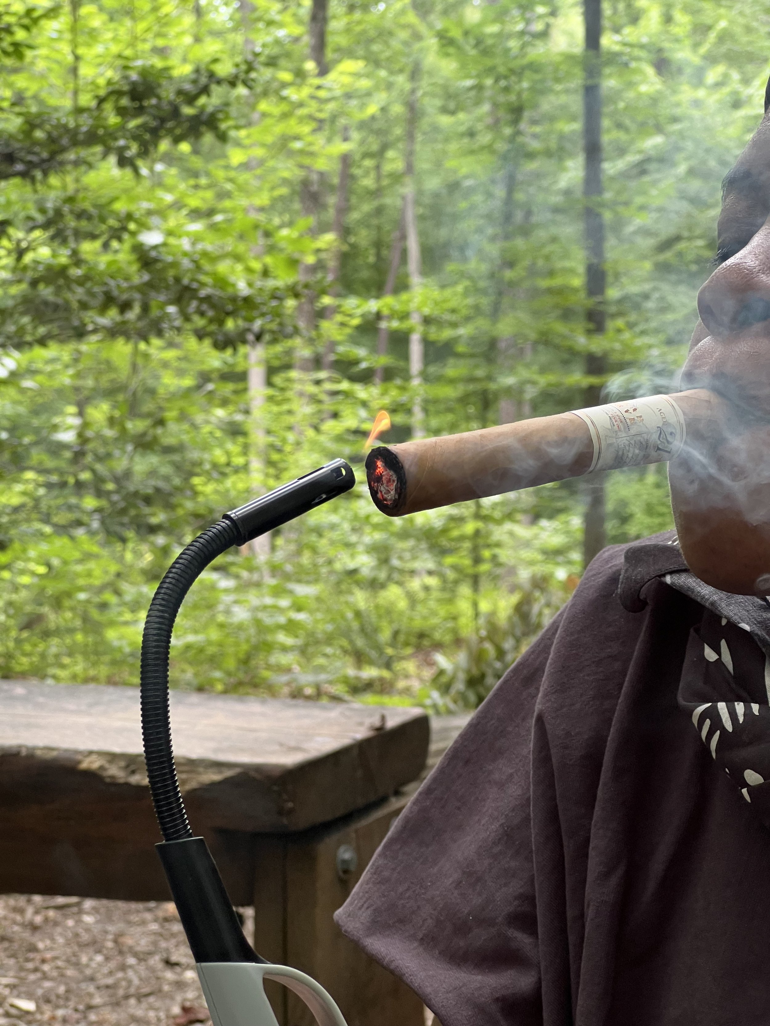 A person smoking a cigar outdoors in a wooded area, with a small torch-like device held near the cigar, creating a visible flame.