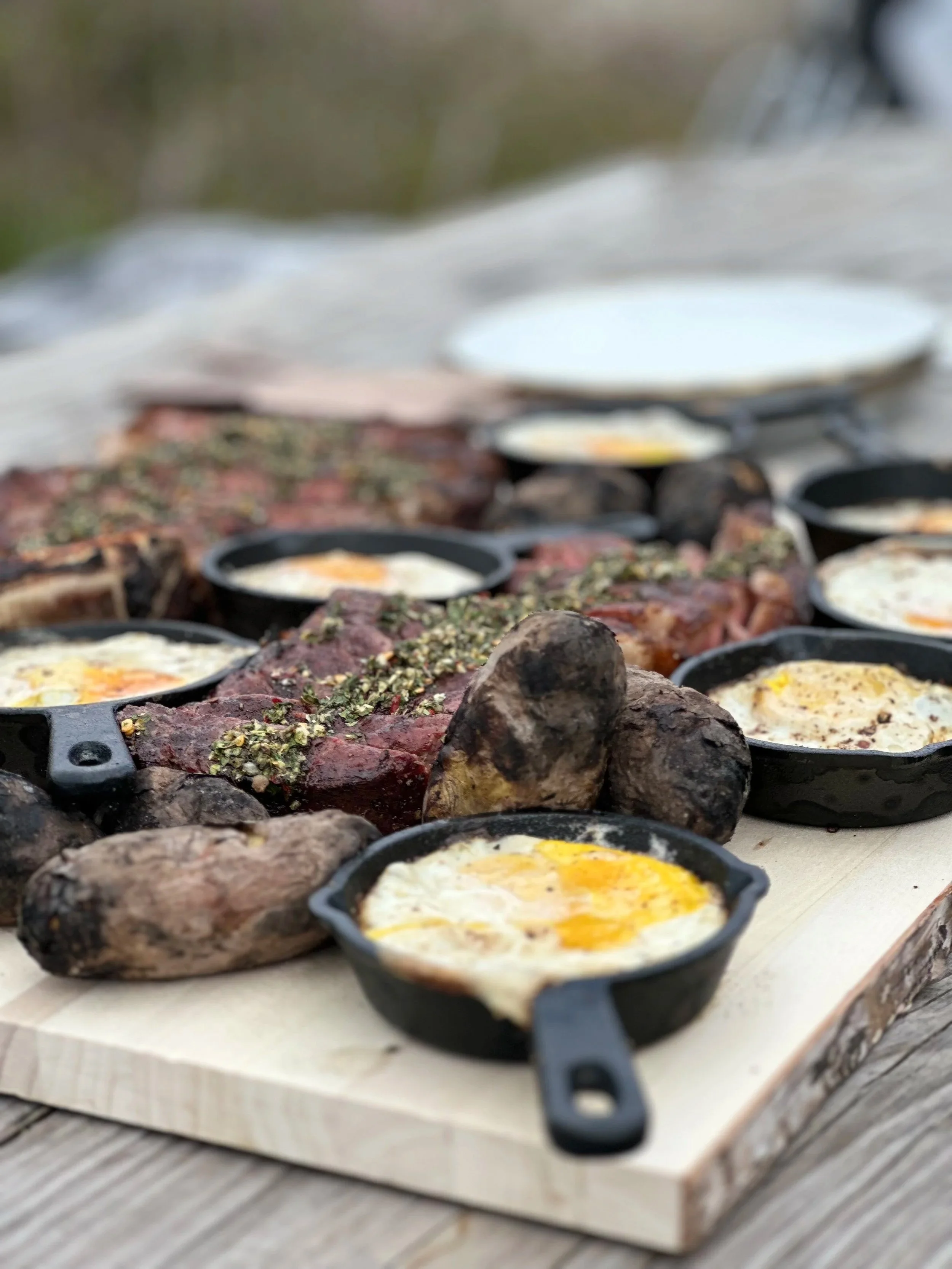 Cast iron skillets with fried eggs, surrounded by cooked meat and topped with herbs on a wooden surface outdoors.