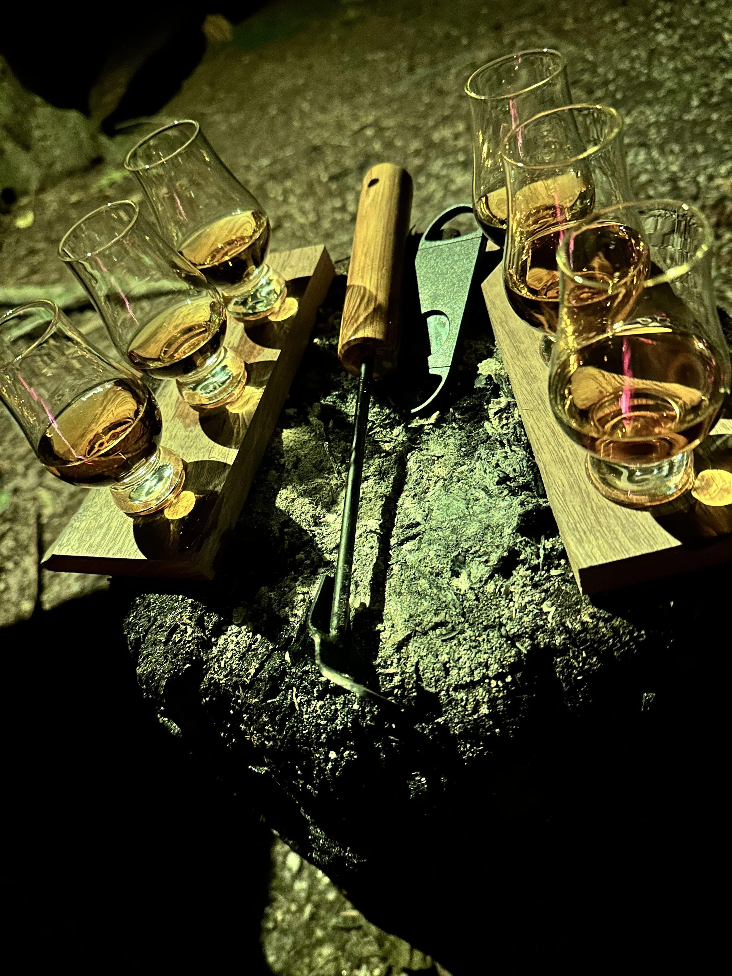 Six glasses of a pinkish beverage on wooden trays, a cheese knife, and a lighter on a rough, cracked surface outdoors.