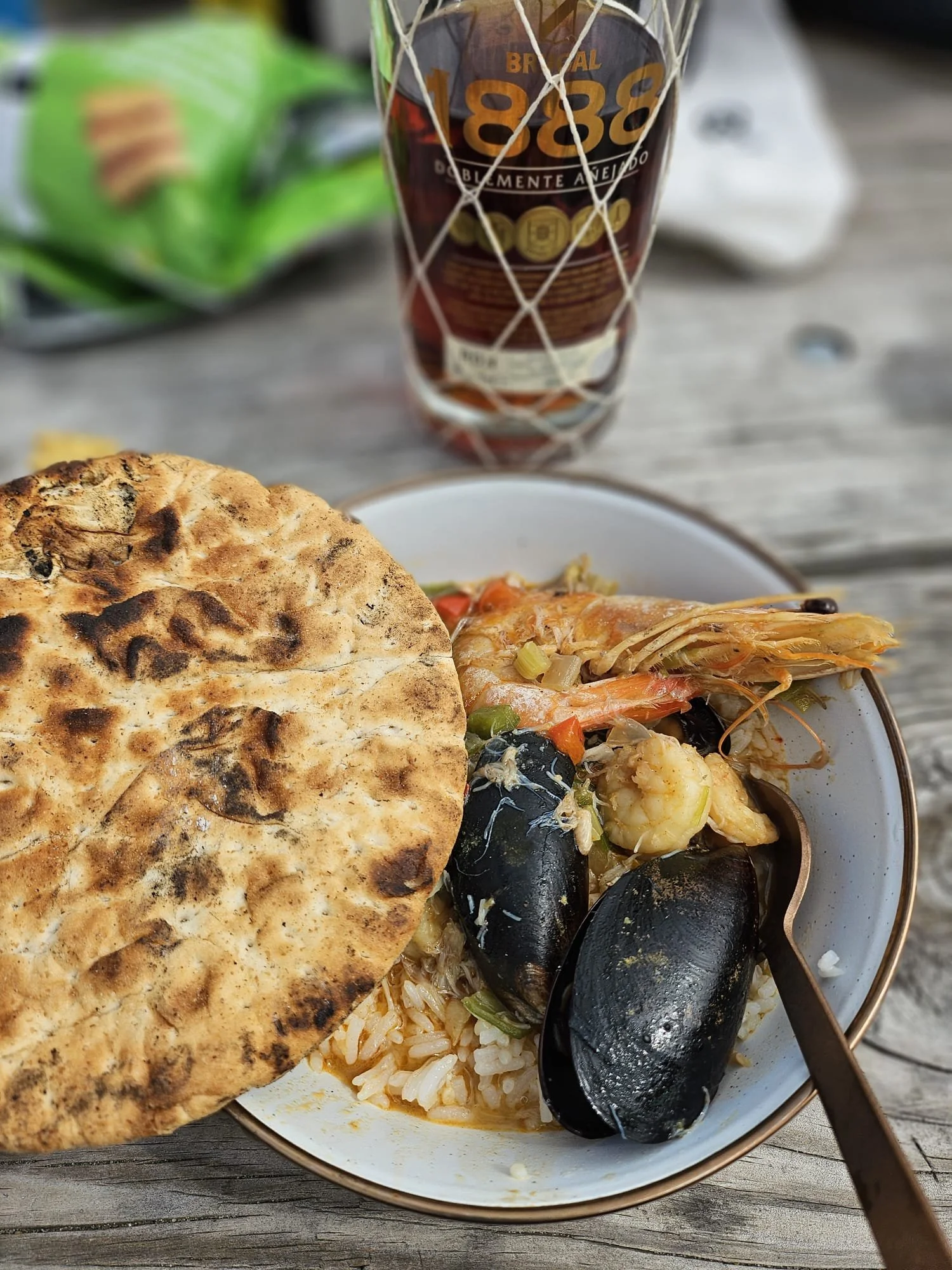 A bowl of seafood with rice, mussels, shrimp, and vegetables, with a piece of naan bread on top, on a wooden table. In the background, there is a glass of beer and a snack bag.
