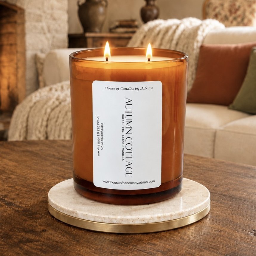 A large amber glass candle jar with two wicks burning, placed on a marble and gold-trimmed round tray on a wooden table. The jar has a white label with black text, indicating it is a candle from House of Candles by Adrian, labeled "Autumn Not Age." The background features a cozy living room with a sofa, throw pillows, and decorative vases.