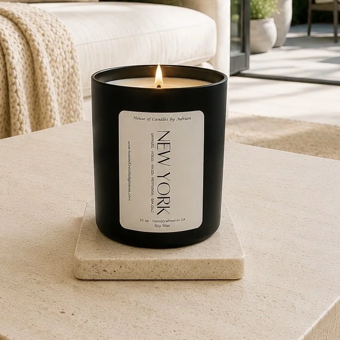 A lit black candle with a white label, placed on a square beige coaster on a wooden table in a cozy living room with a couch, blanket, and plants in the background.