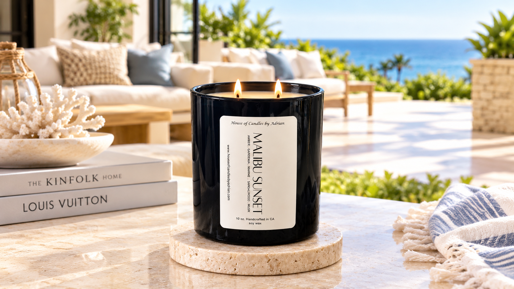 A black candle labeled 'Malibu Sunset' on a beige marble stand on a light-colored marble table, with a bright outdoor patio and ocean view in the background, decorated with pillows and tropical plants.