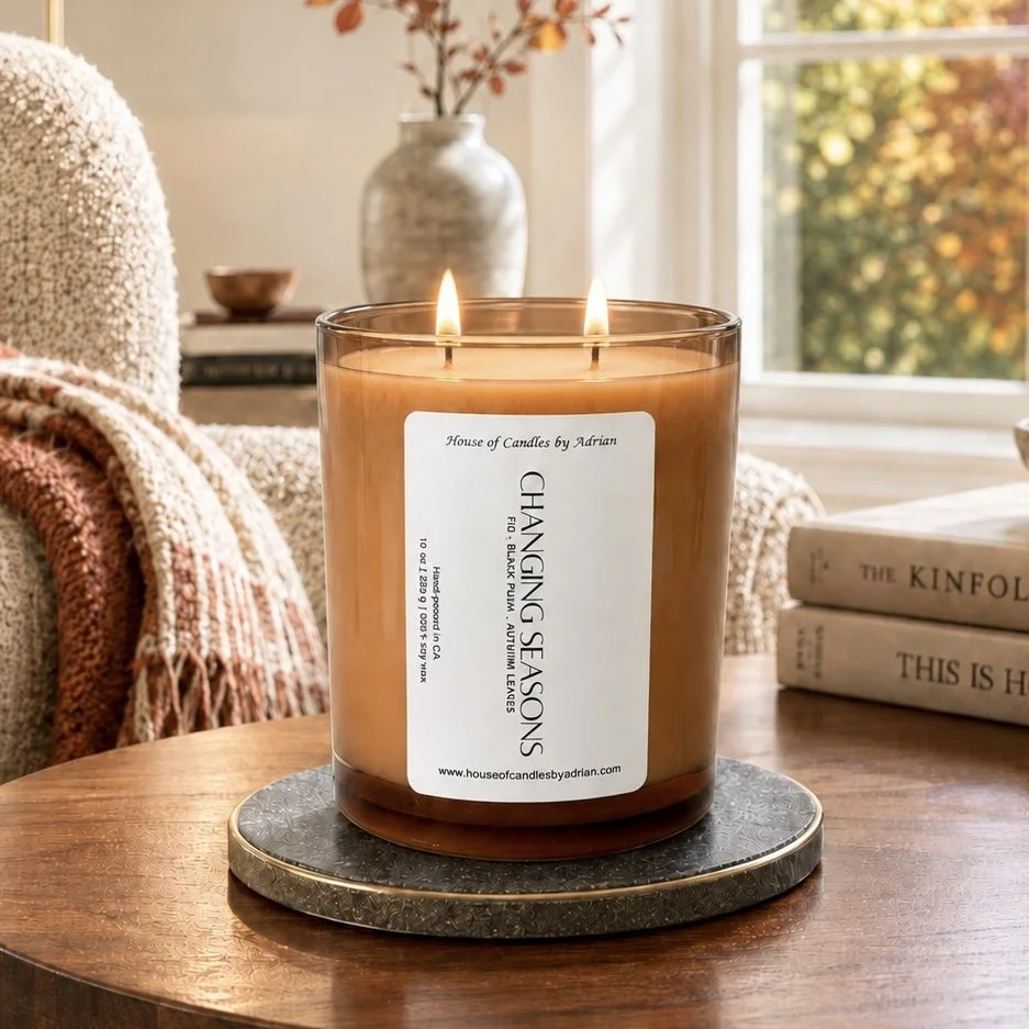 A large brown candle with two wicks burning on a wooden table, labeled 'Chasing Seasons' from House of Candles by Adrian. The candle is placed on a round black and gold tray. In the background, there is a cozy setting with a beige upholstered chair, a blanket, a vase with branches, and a window showing autumn trees.