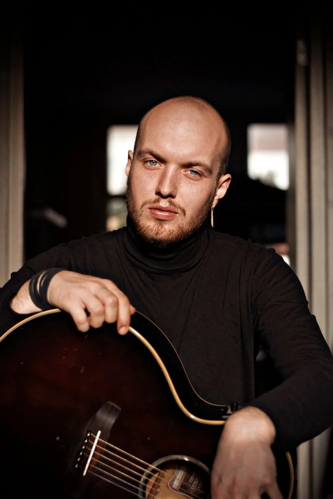 Portret van een man met in een zwart shirt, die een gitaar vasthoudt, binnen. Donker achtergrond met enkele lichte raampjes.