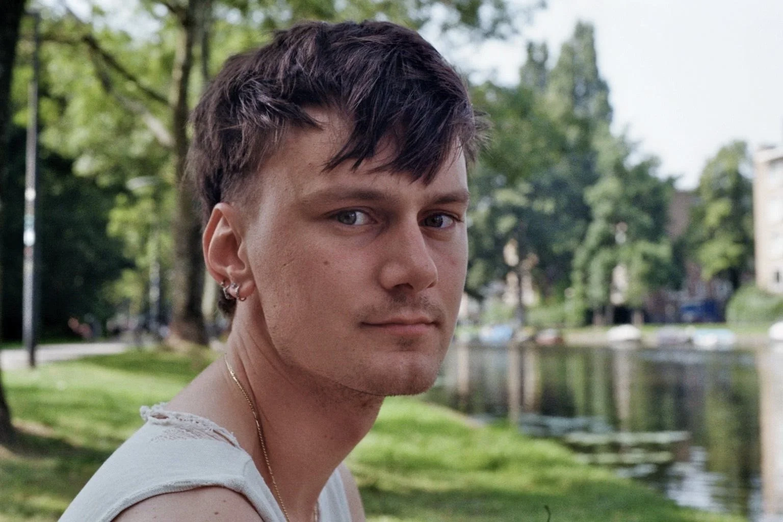 Portret van een jonge man met donkerbruin haar met een plukje naar voren, blauwe ogen, lichte huid met een paar sproeten, die naar de camera kijkt. Hij draagt een witte top met versleten randjes, oorbellen en een ketting, met een groene park, bomen en een waterpartij op de achtergrond.