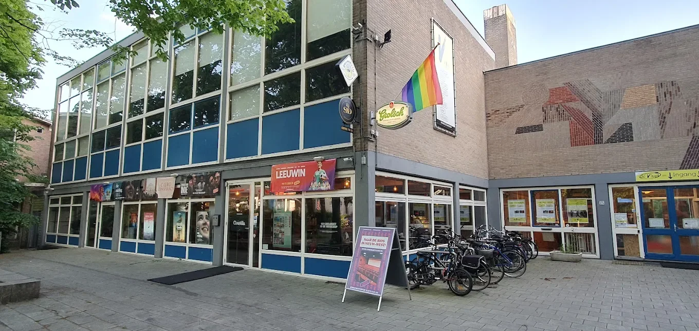 Een gebouw met grote ramen en vlaggen, waaronder een regenboogvlag, en fietsen voor de ingang.