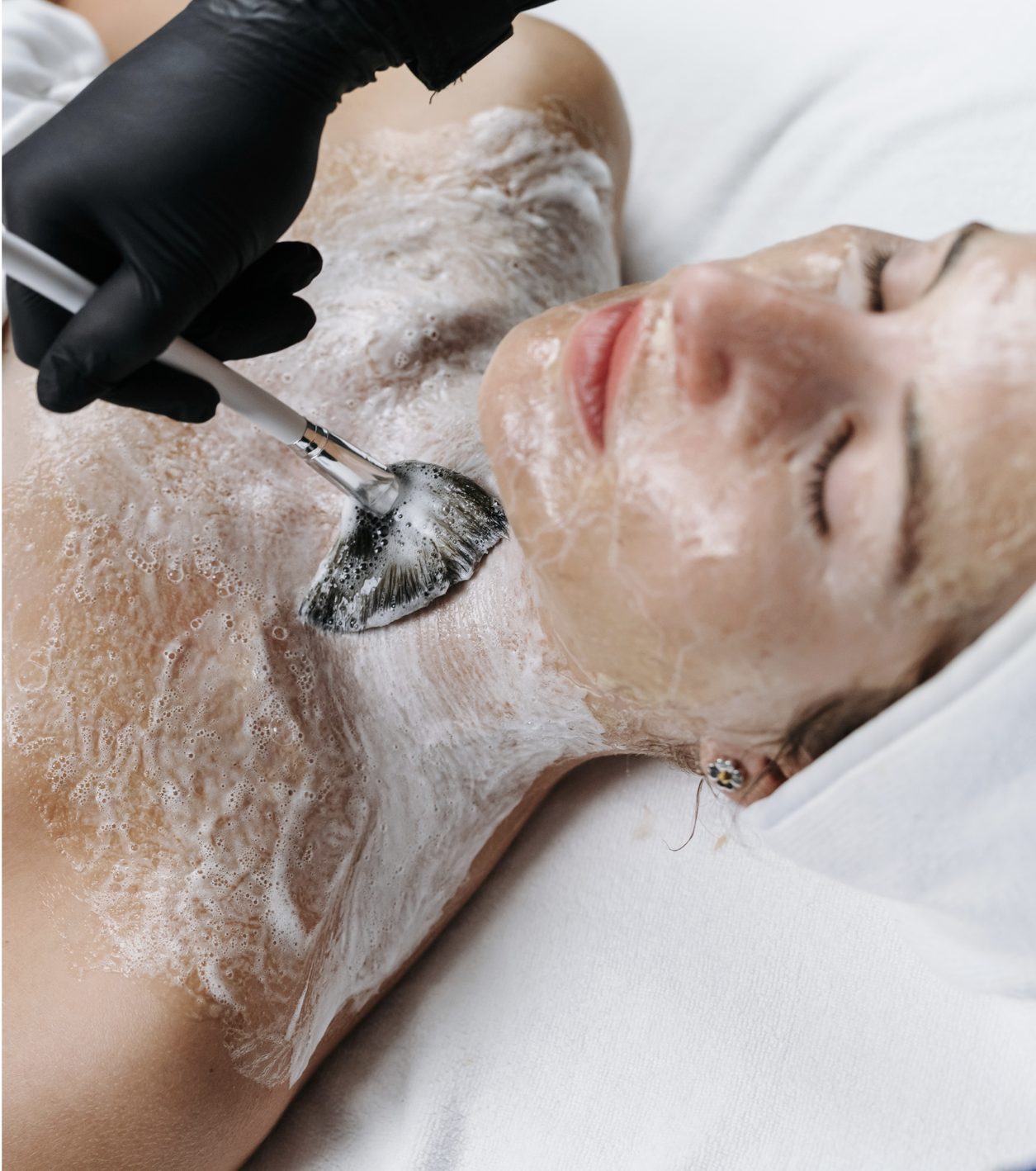 A woman receiving a facial treatment with a brush application of foam or cleansing product on her face and chest, lying down with eyes closed.