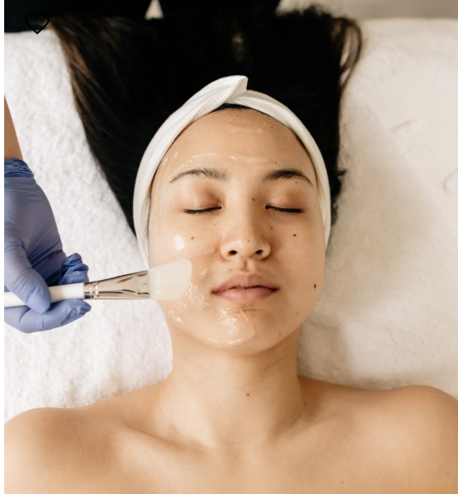 A woman lying back with eyes closed, receiving a facial treatment with a brush on her cheek, wearing a headband, in a spa setting.