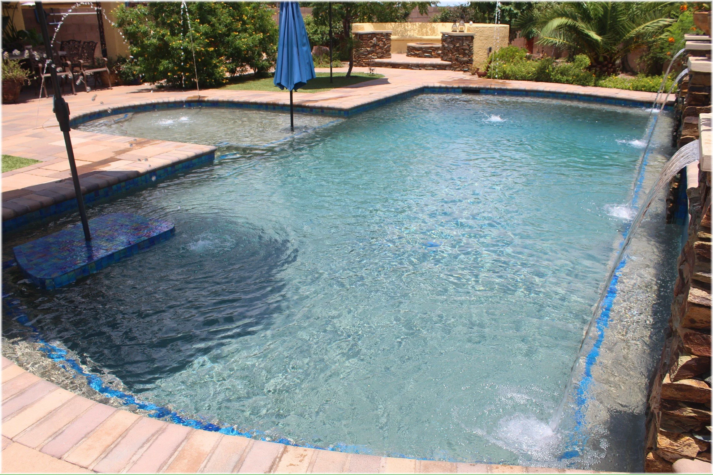 Freeform custom swimming pool with a tanning ledge, blue water, and travertine deck, designed and built by Meraki Pools AZ in Tucson, Arizona.