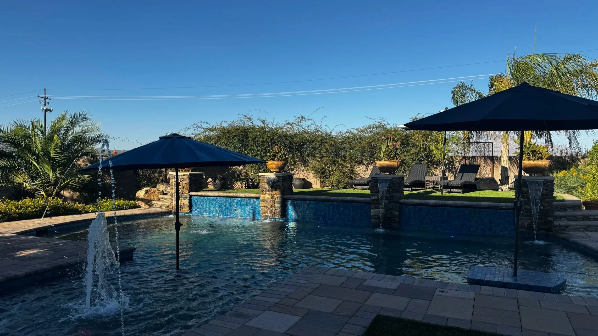Backyard pool area with blue umbrellas, lounge chairs, and a water fountain, surrounded by trees and greenery under a clear blue sky.