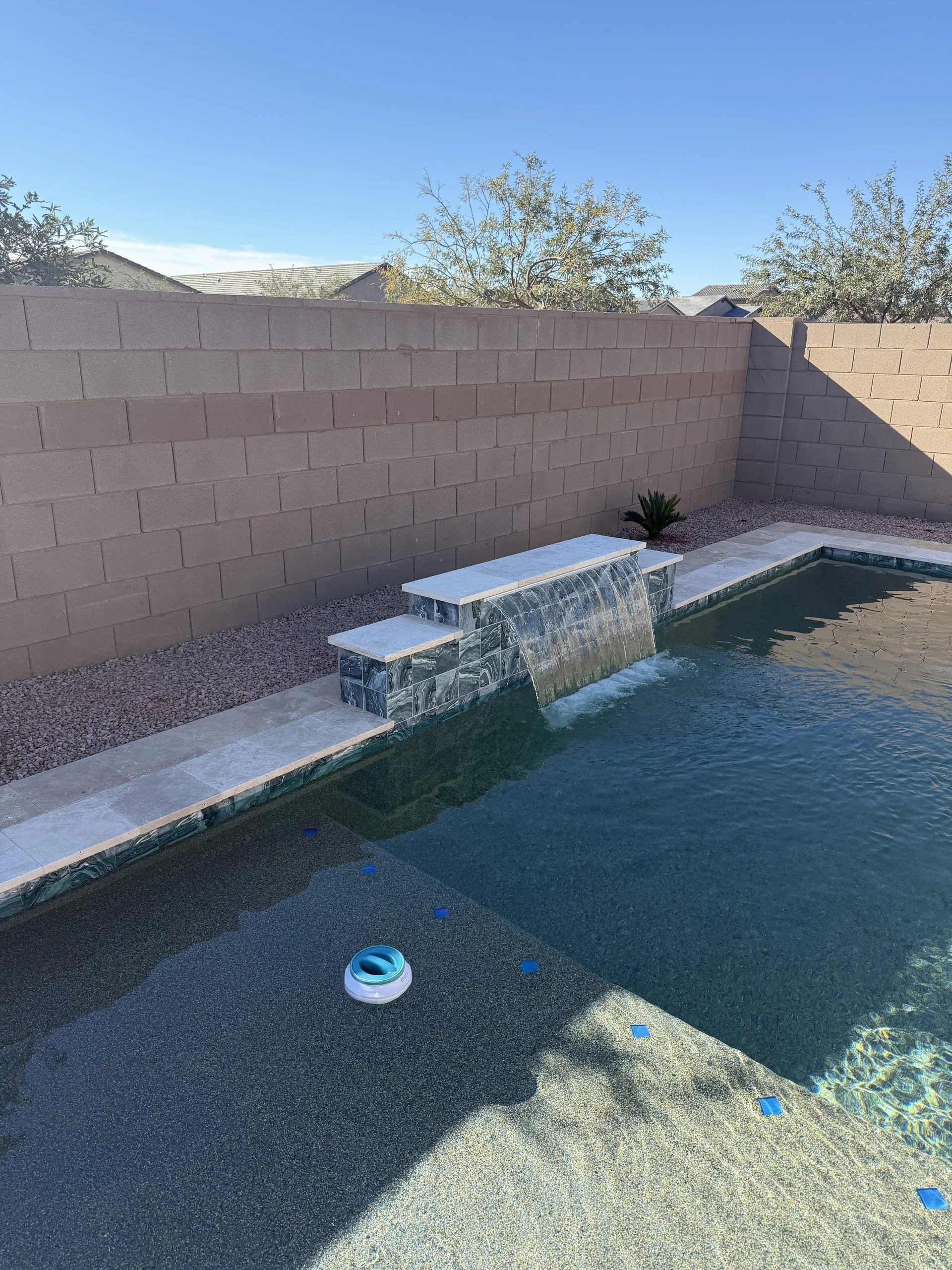 Custom pool with a sheer-descent water feature spilling into blue water, framed by travertine decking, by Meraki Pools AZ in Tucson, Arizona.