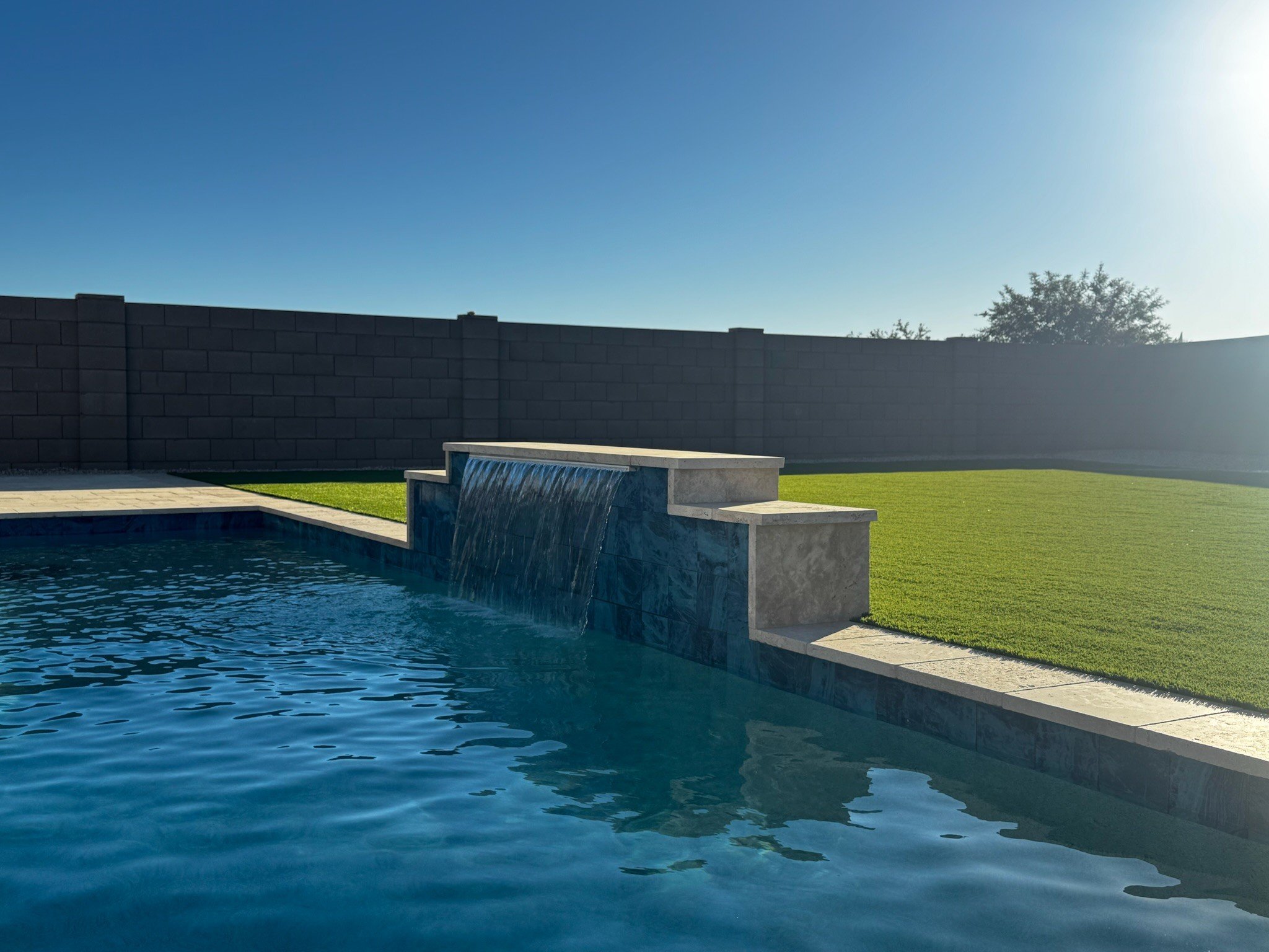 A swimming pool with blue water, a water feature on the edge, a tall dark brick wall in the background, and a lush green lawn with a tree and clear blue sky above.