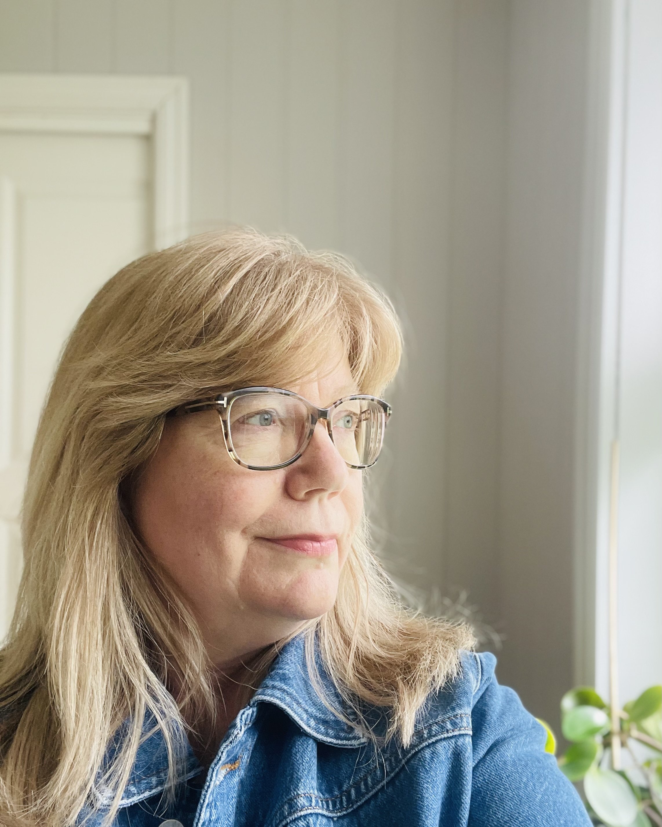 En kvinne med blonde hår og briller ser ut av et vindu, iført en blå denimjakke, med grønne planter i bakgrunnen.