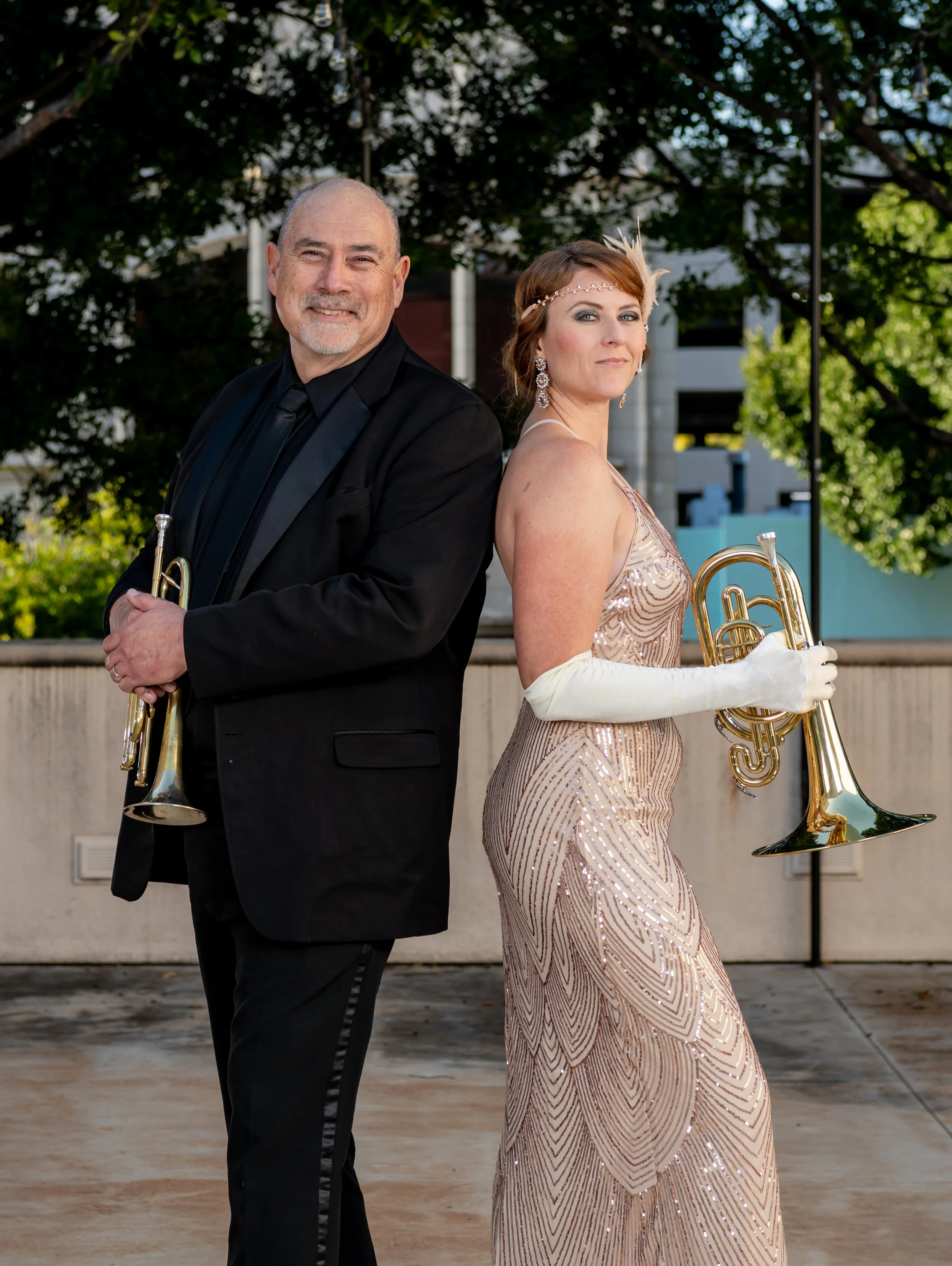 A man in a black tuxedo and a woman in a glamorous, sequined dress holding brass instruments, standing outdoors with trees and buildings in the background.