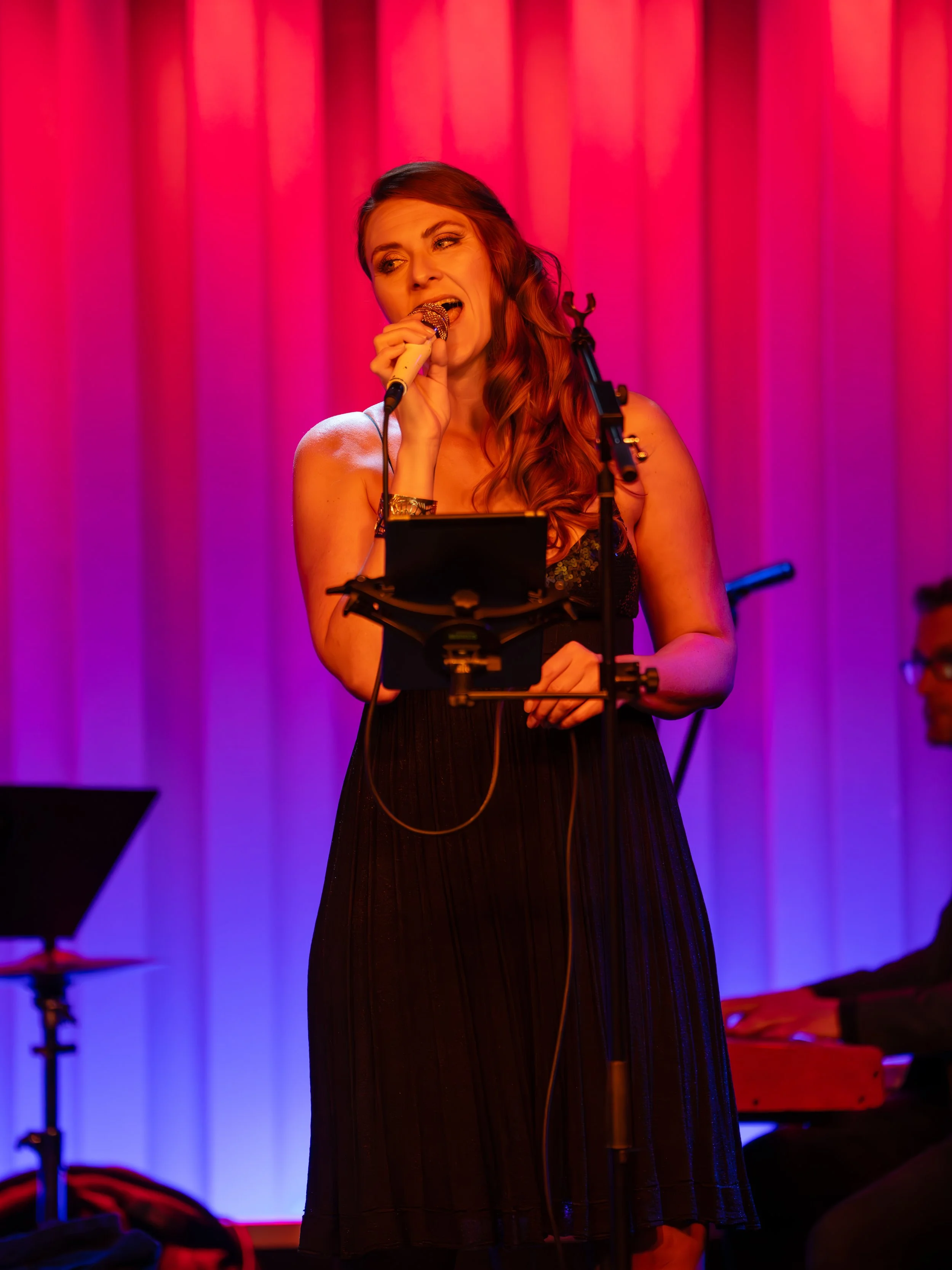 Female singer with long wavy hair performing on stage with purple and pink lighting, holding a microphone, and a musician playing keyboard in the background.