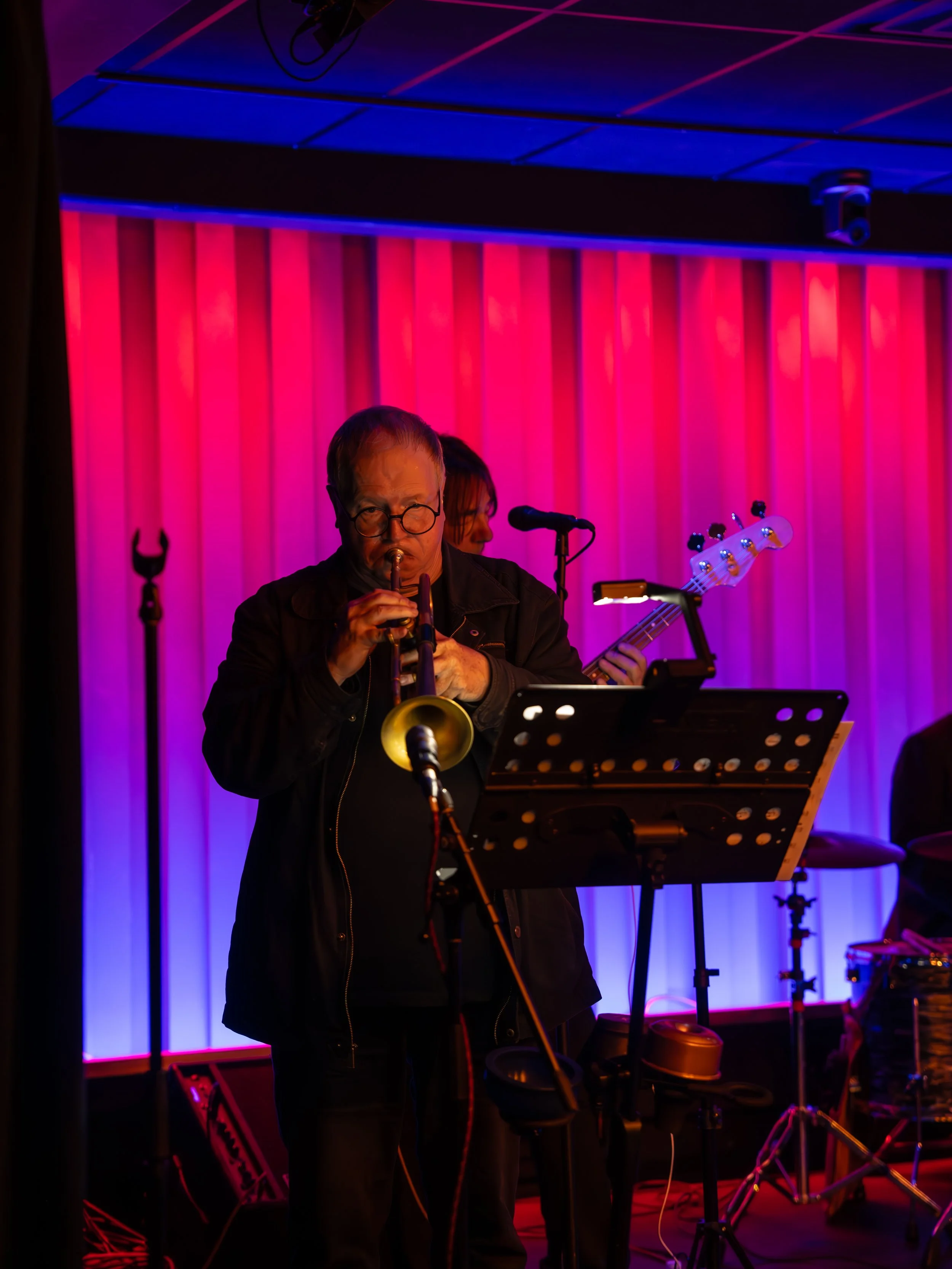 A musician playing a trumpet on stage with colorful pink and blue lighting in the background.