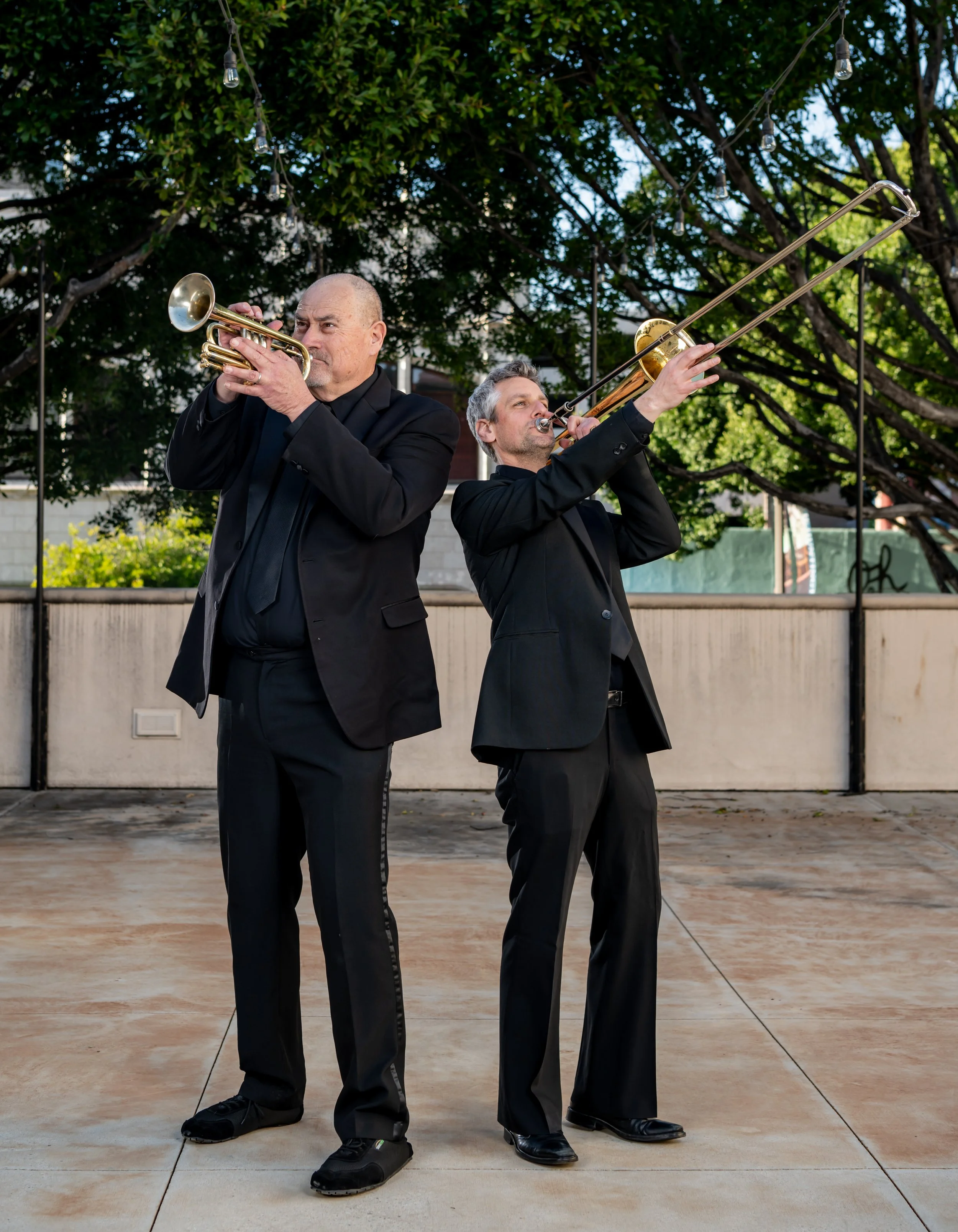 Two men in black suits playing brass instruments, one with a trumpet and the other with a trombone, outdoors under string lights and trees.