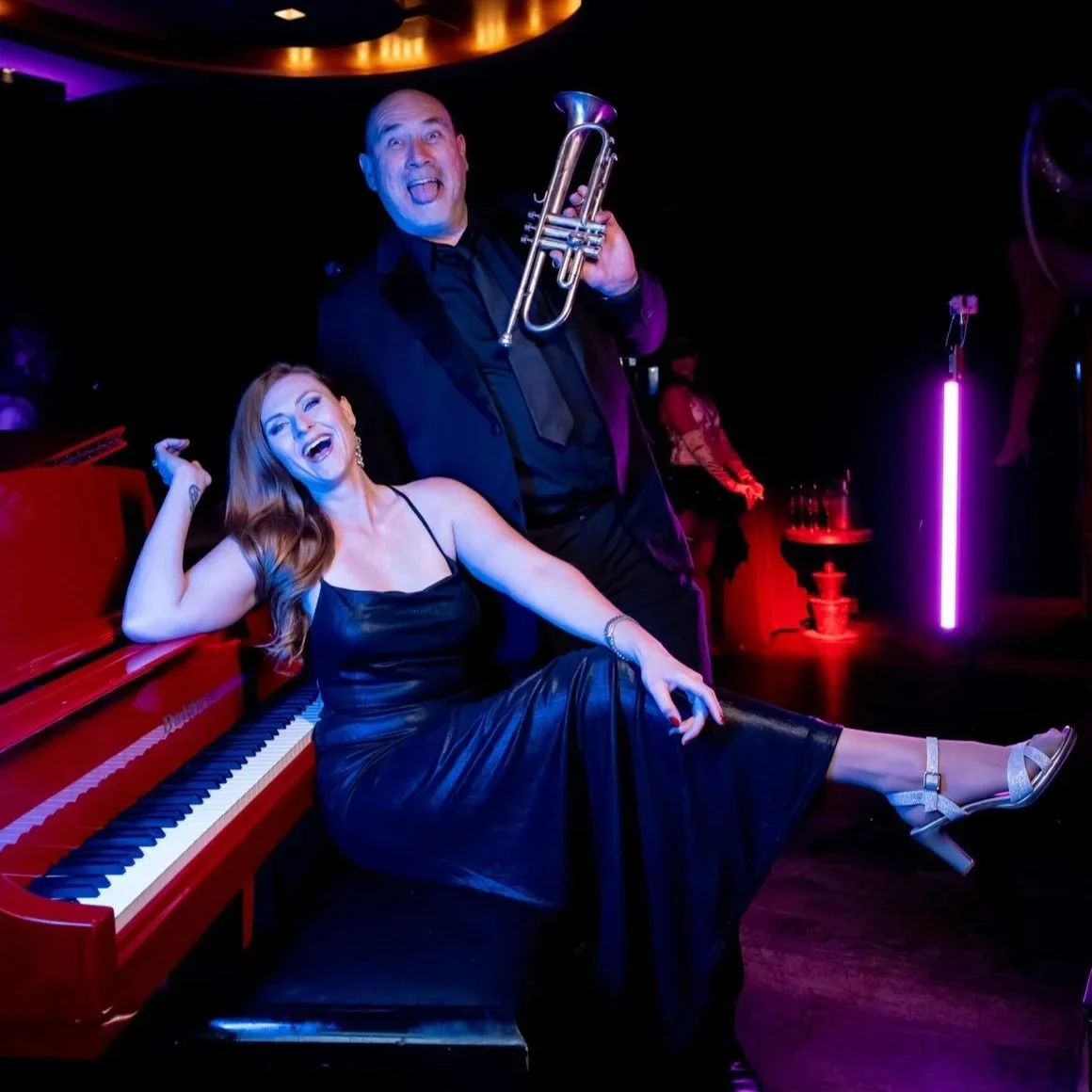 A woman sitting on a red piano with a man standing behind her holding a trumpet in a dimly lit elegant nightclub or lounge. Both are smiling and appear joyful, dressed in formal attire.