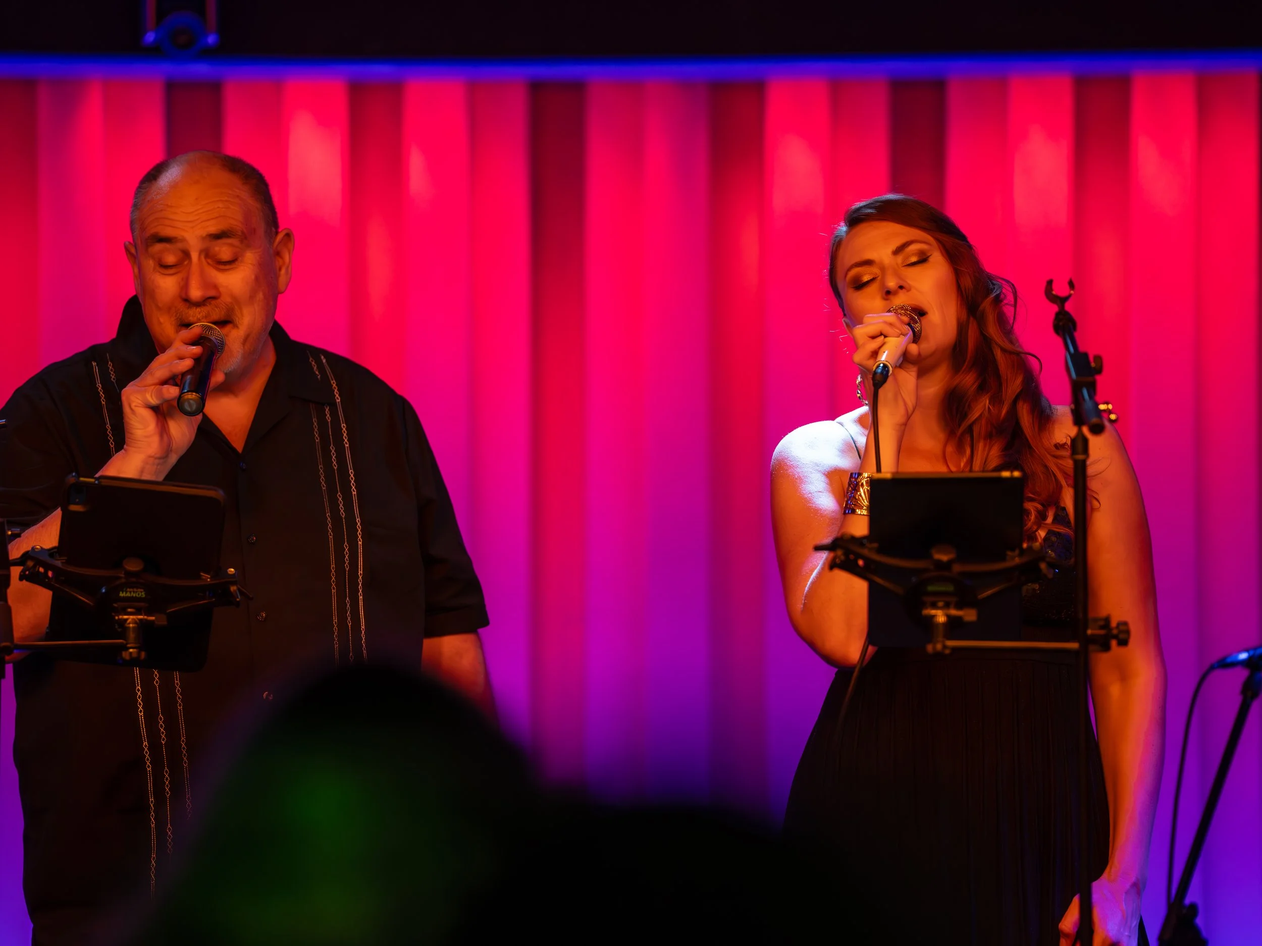 Two singers performing on stage under pink and purple lighting with a pink curtain background.
