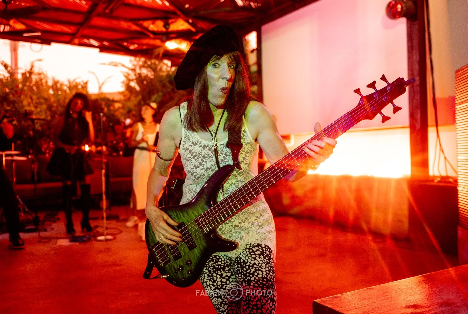 A woman playing an electric bass guitar at an indoor event with warm, orange lighting. She is wearing a black hat, a sleeveless lace top, and patterned pants. Background shows other people and some equipment.
