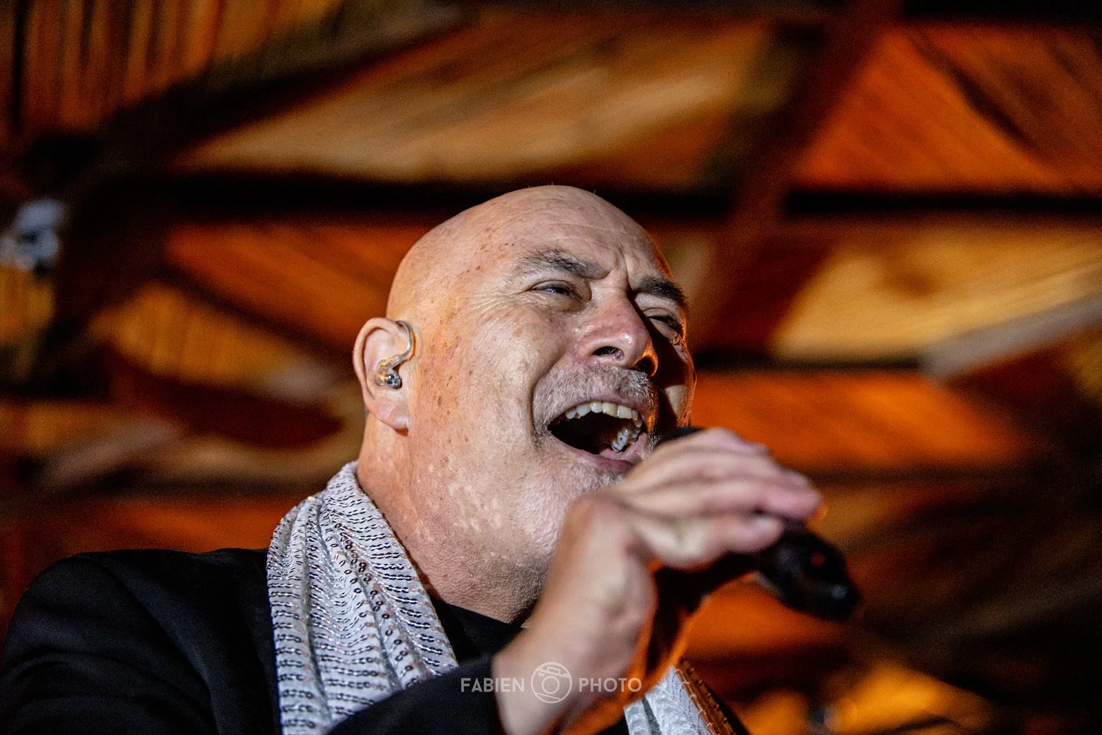 A bald man with a mustache and goatee singing into a microphone, wearing a silver scarf and in-ear monitors, against a warm, blurred wooden background.
