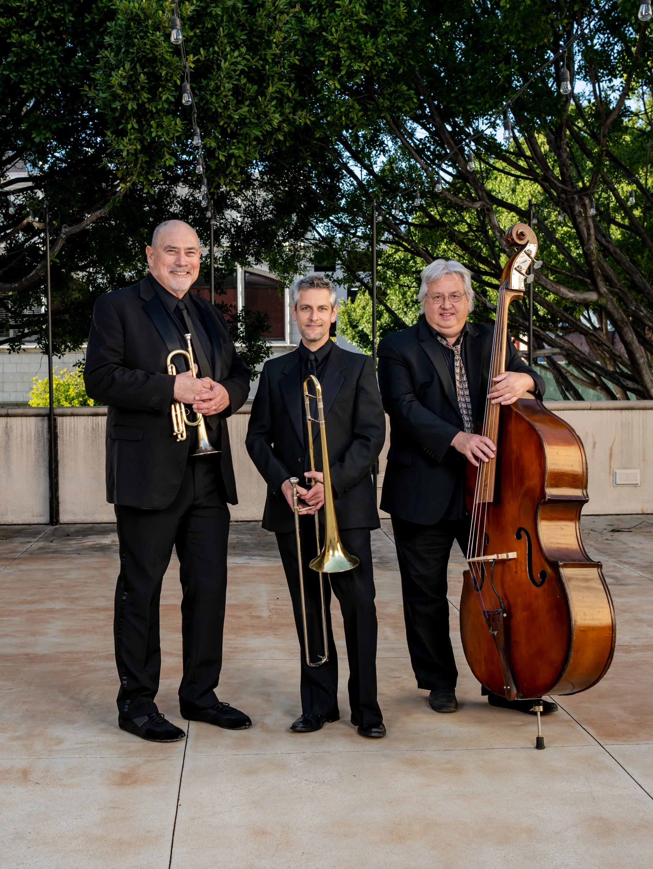 Three male jazz musicians in black suits with instruments: trumpet, trombone, and double bass, standing outdoors on a concrete patio with trees and string lights.