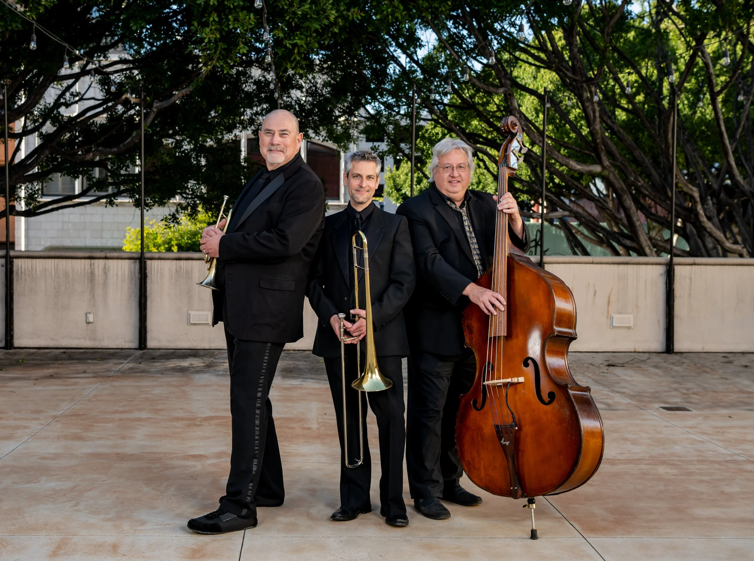 Three men in black suits standing outdoors, each holding a musical instrument: trumpet, trombone, and double bass, with trees and a building in the background.