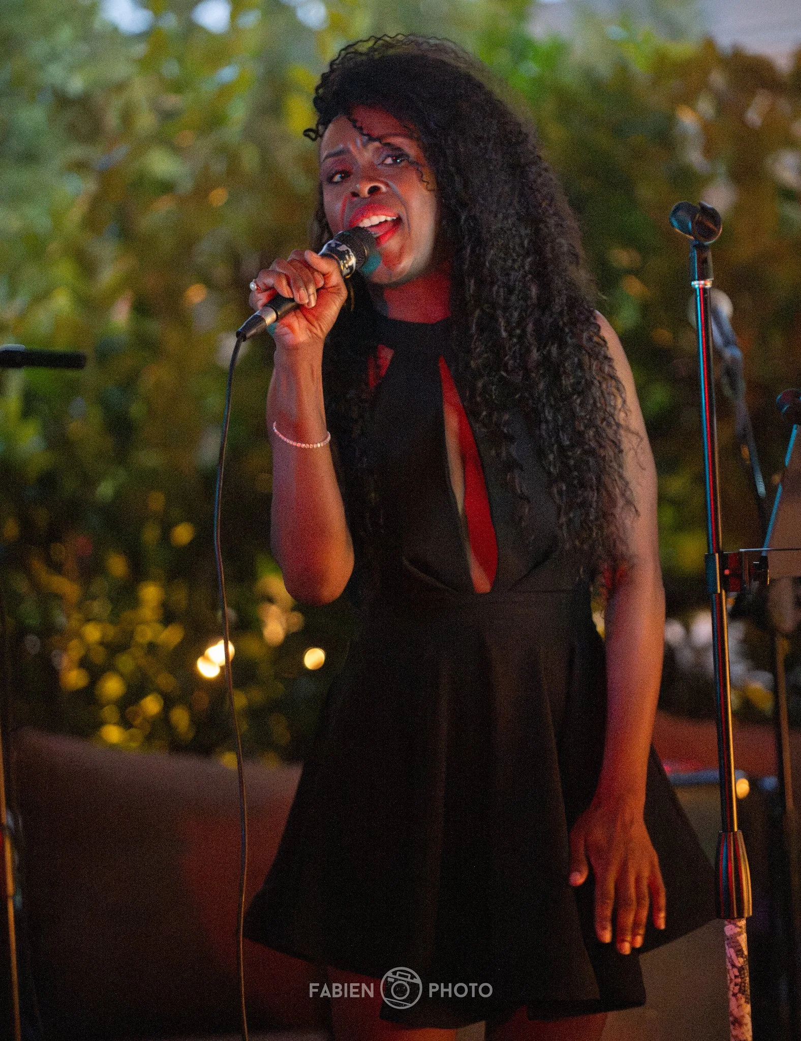 A woman with long curly black hair sings into a microphone at an outdoor event during the evening, wearing a black dress with a red accent.