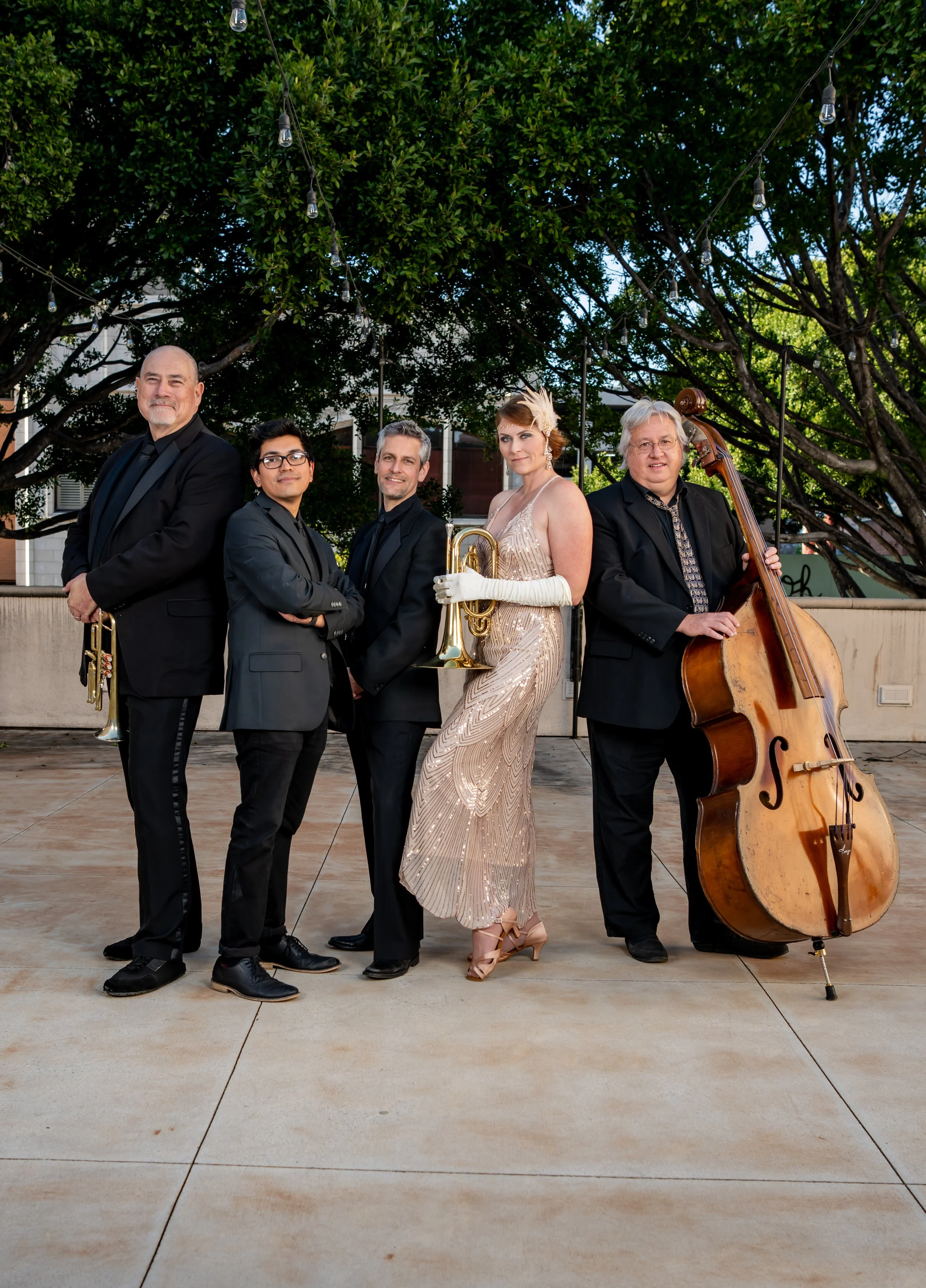 A jazz band performing outdoors under string lights, featuring five musicians with a mix of instruments including a trumpet, double bass, and wind instruments. They are dressed in formal black attire, with a woman in a vintage-style gown holding a tr
